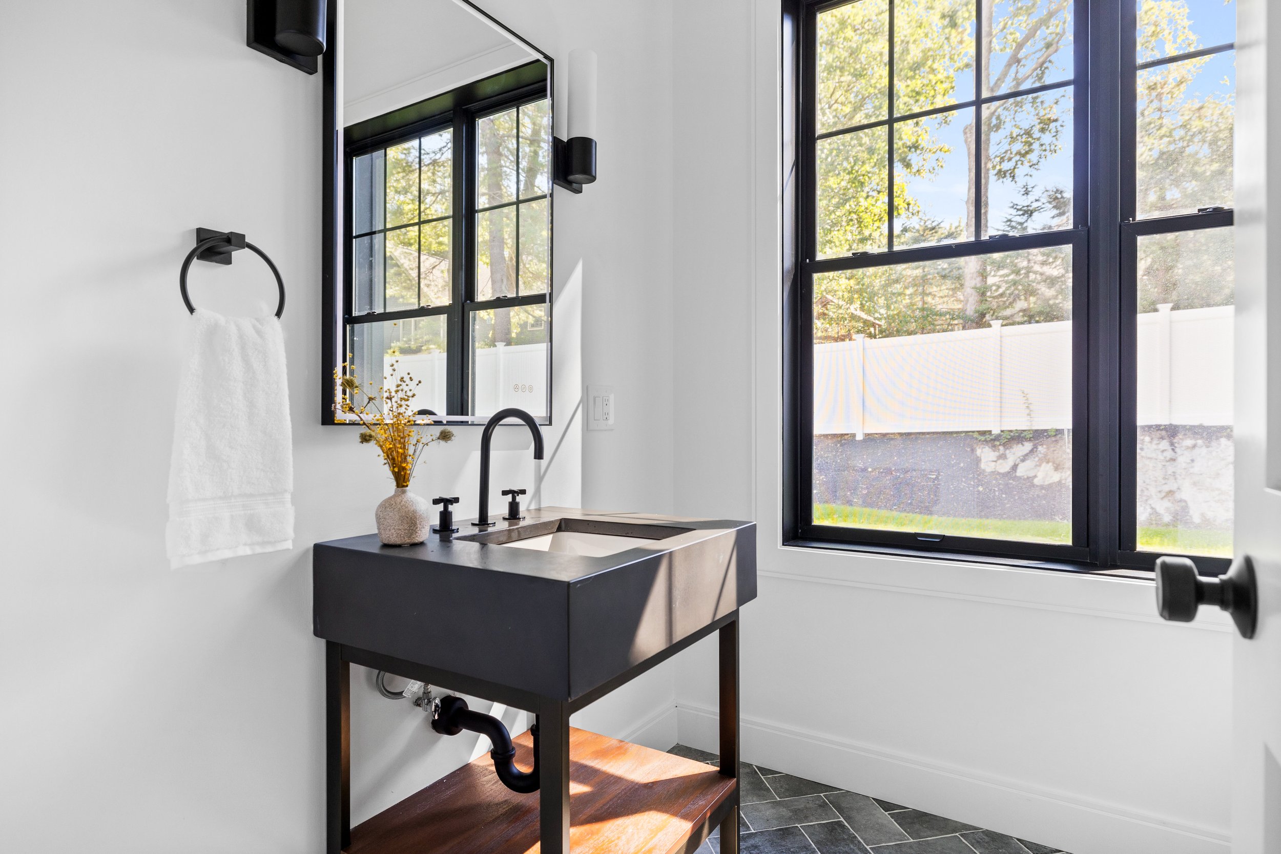 Modern laundry room with black utility sink, white walls, large window with black frame showing outdoor trees, and a small vase with dried flowers on the sink