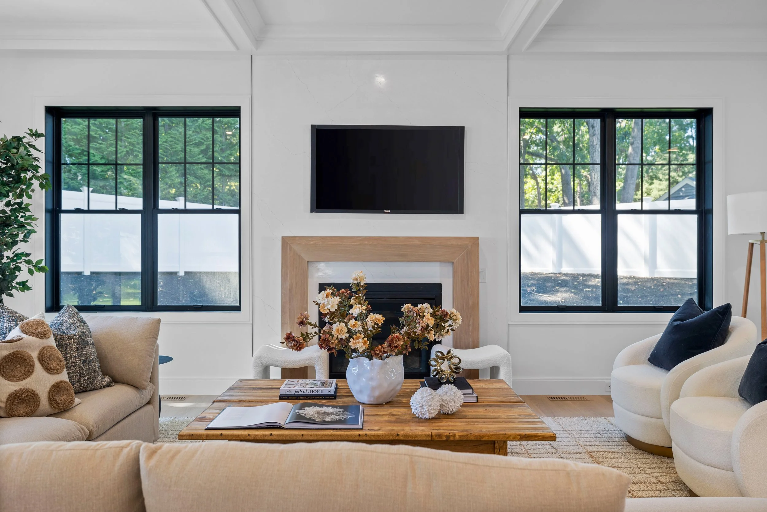 Living room with a cream sofa, white armchair, wooden coffee table, floral arrangement, fireplace, black wall-mounted TV, and two large windows with greenery outside.