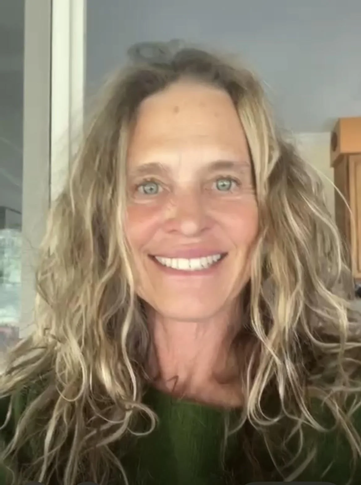A woman with long, curly blonde hair, light skin, and blue eyes smiling at the camera indoors.