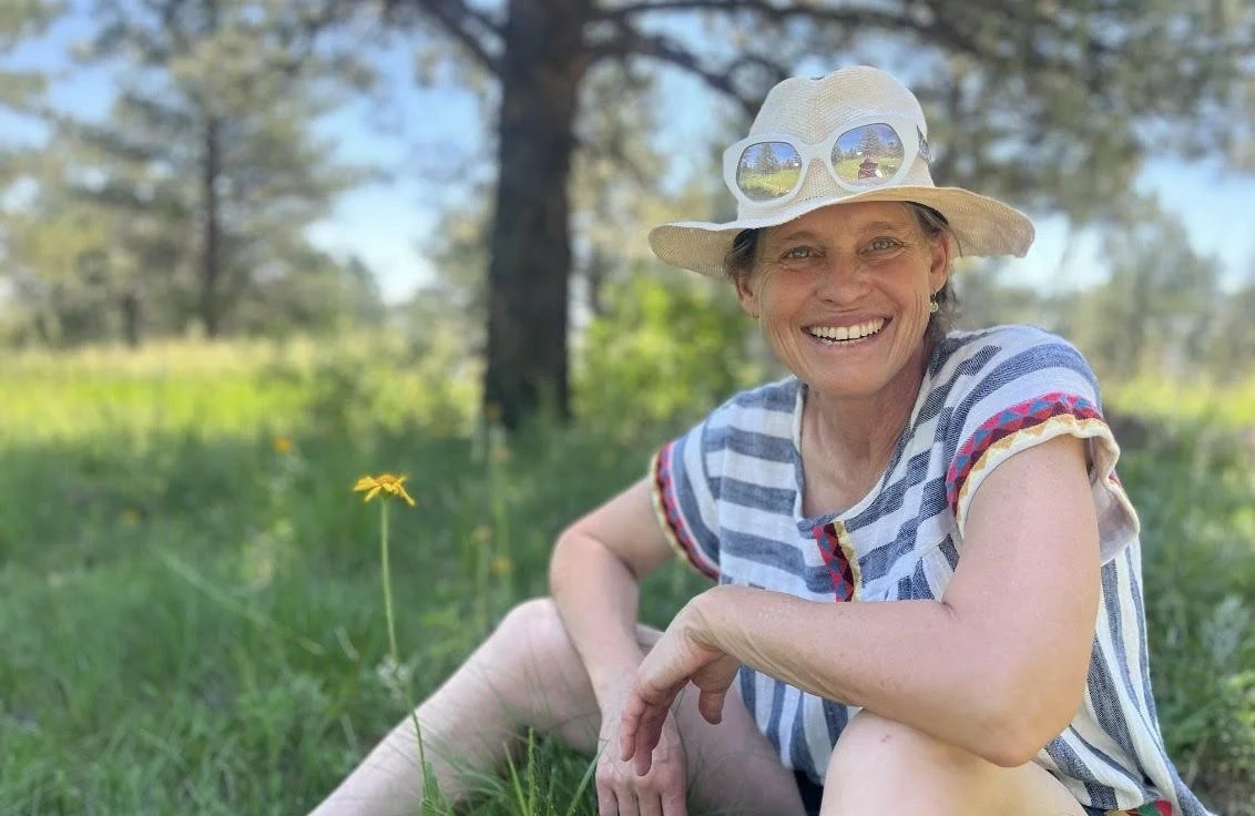 A smiling woman wearing a wide-brim hat with sunglasses sitting outdoors in a grassy area with trees in the background.