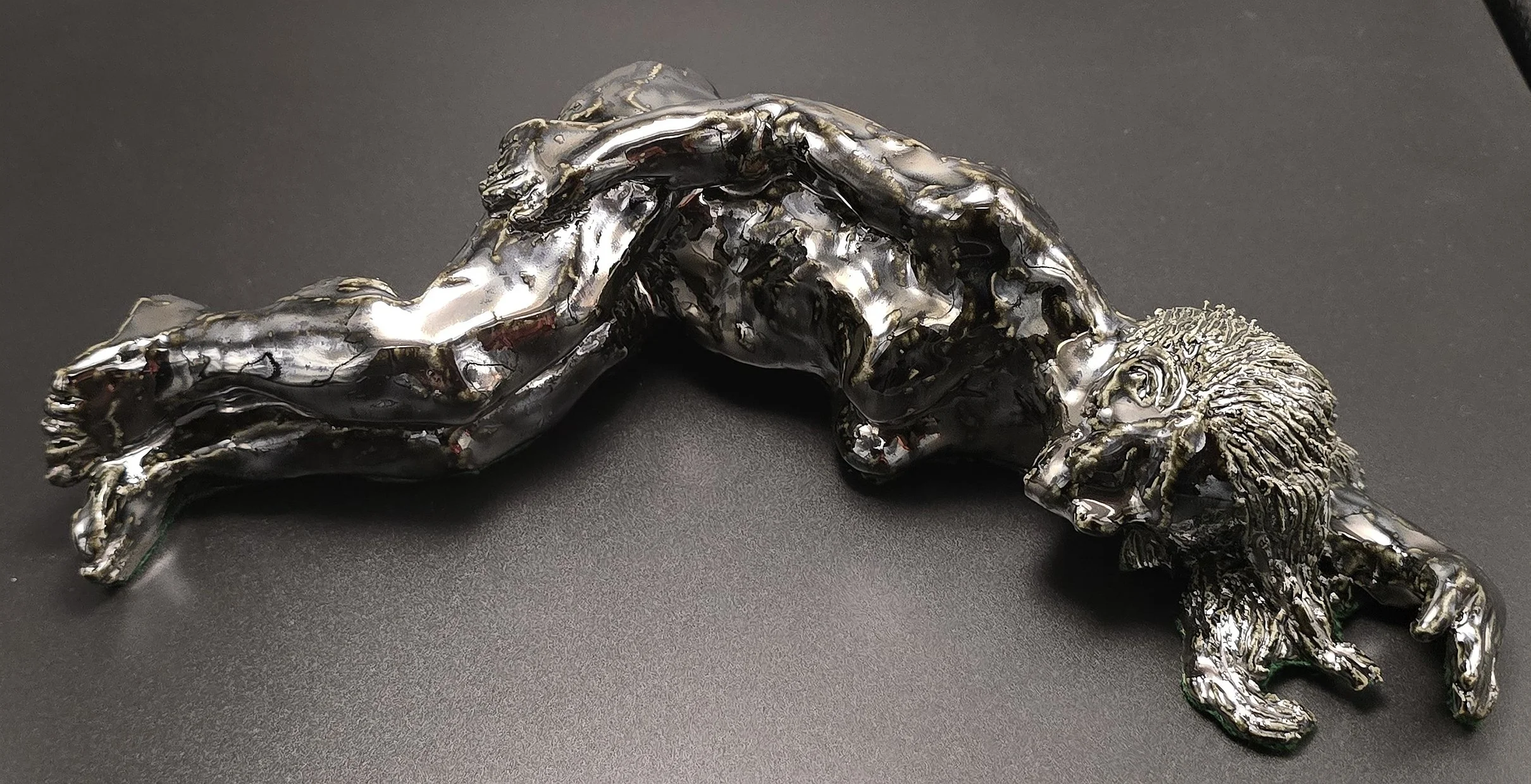 Silver- or palladium-glazed  ceramic sculpture of a reclining woman with long hair, lying on her side with one arm behind her head, on a black background.