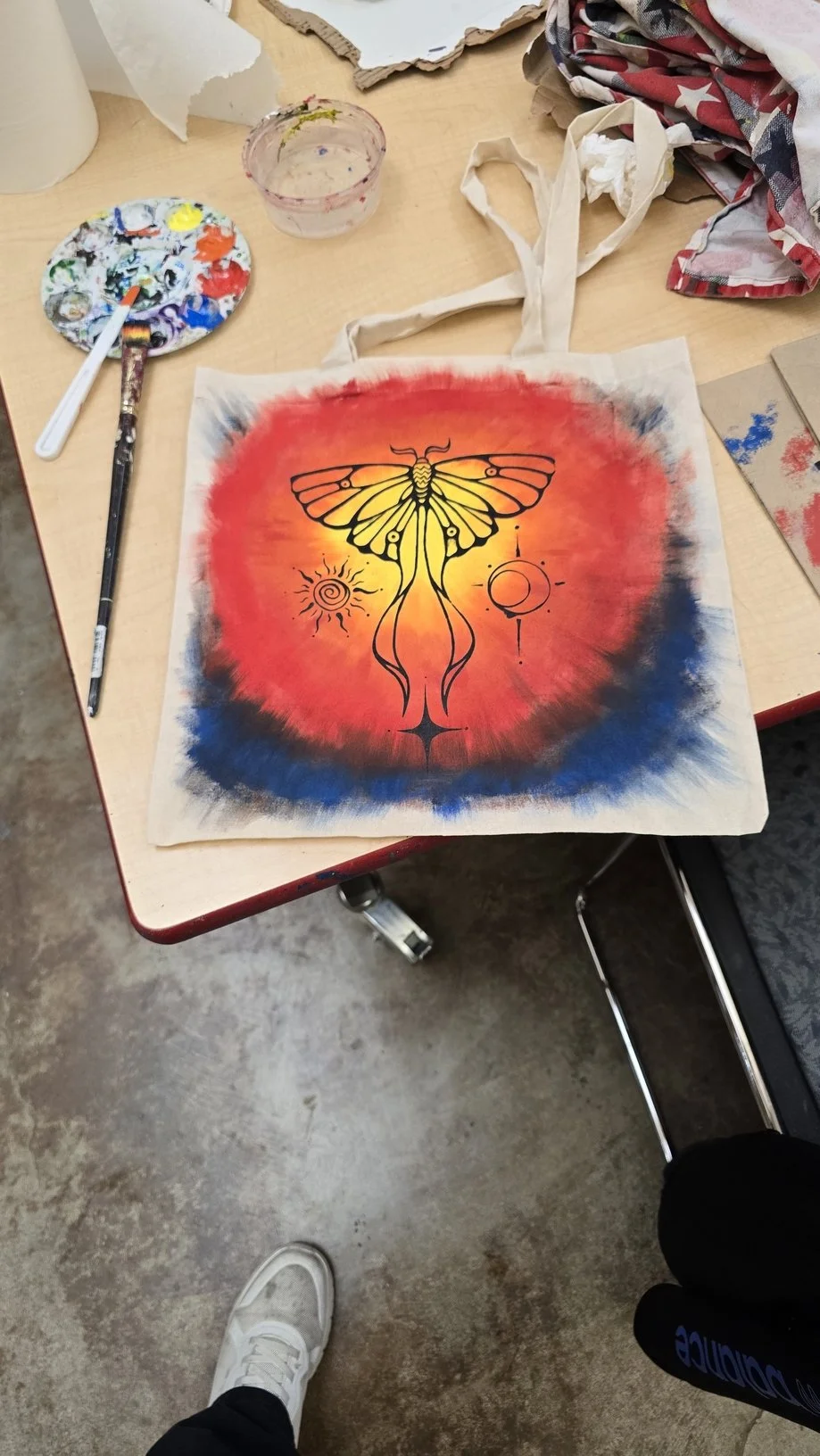 A tote bag with an illustration of a yellow moth against a red and blue abstract background, with symbols resembling the sun and the moon.