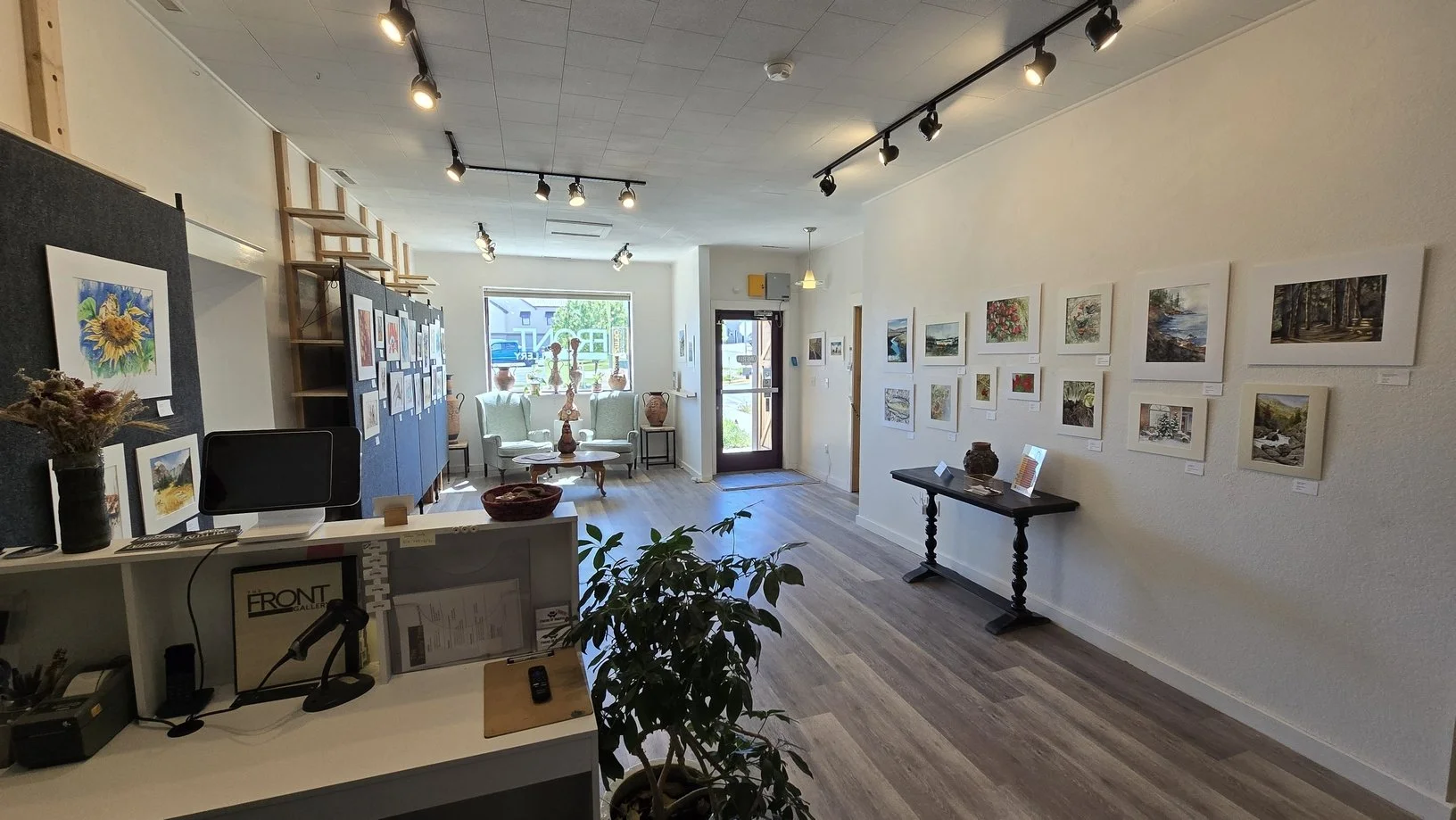 Interior of an art gallery with landscape paintings on the white walls, a reception desk with a computer, and art decorations like vases and sculptures near the window and door.