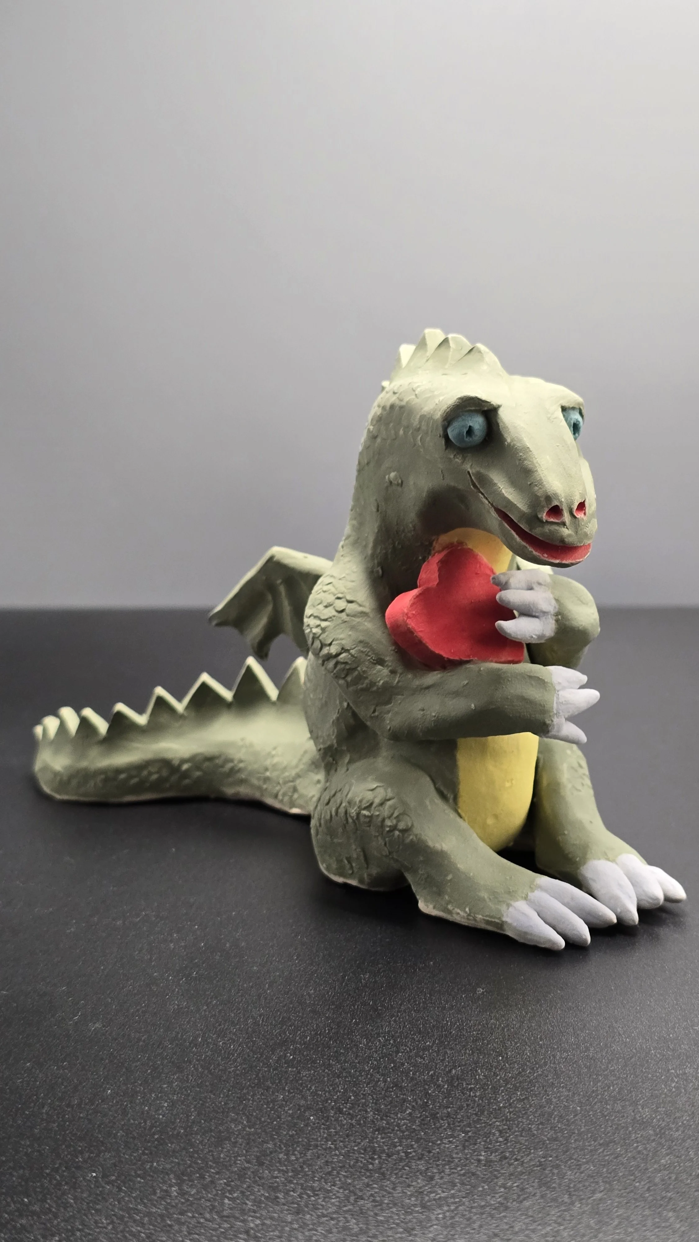 A small ceramic sculpture of a fat happy dragon holding a red heart in its front limbs.