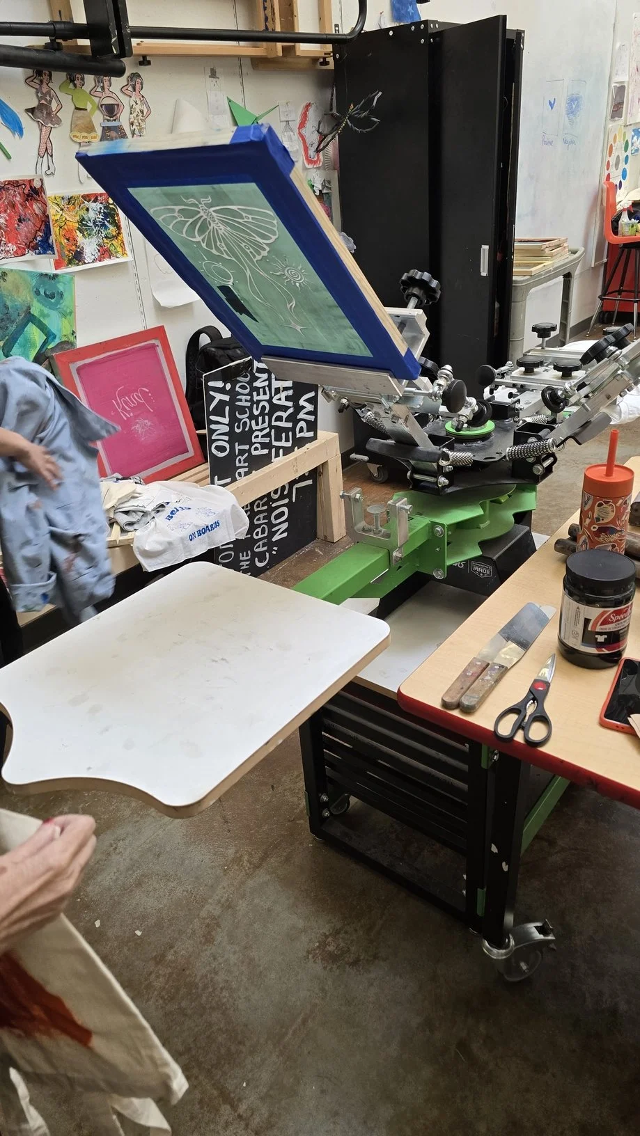A silk-screen printing press in an art studio, with artwork, scissors, and art supplies surrounding it.