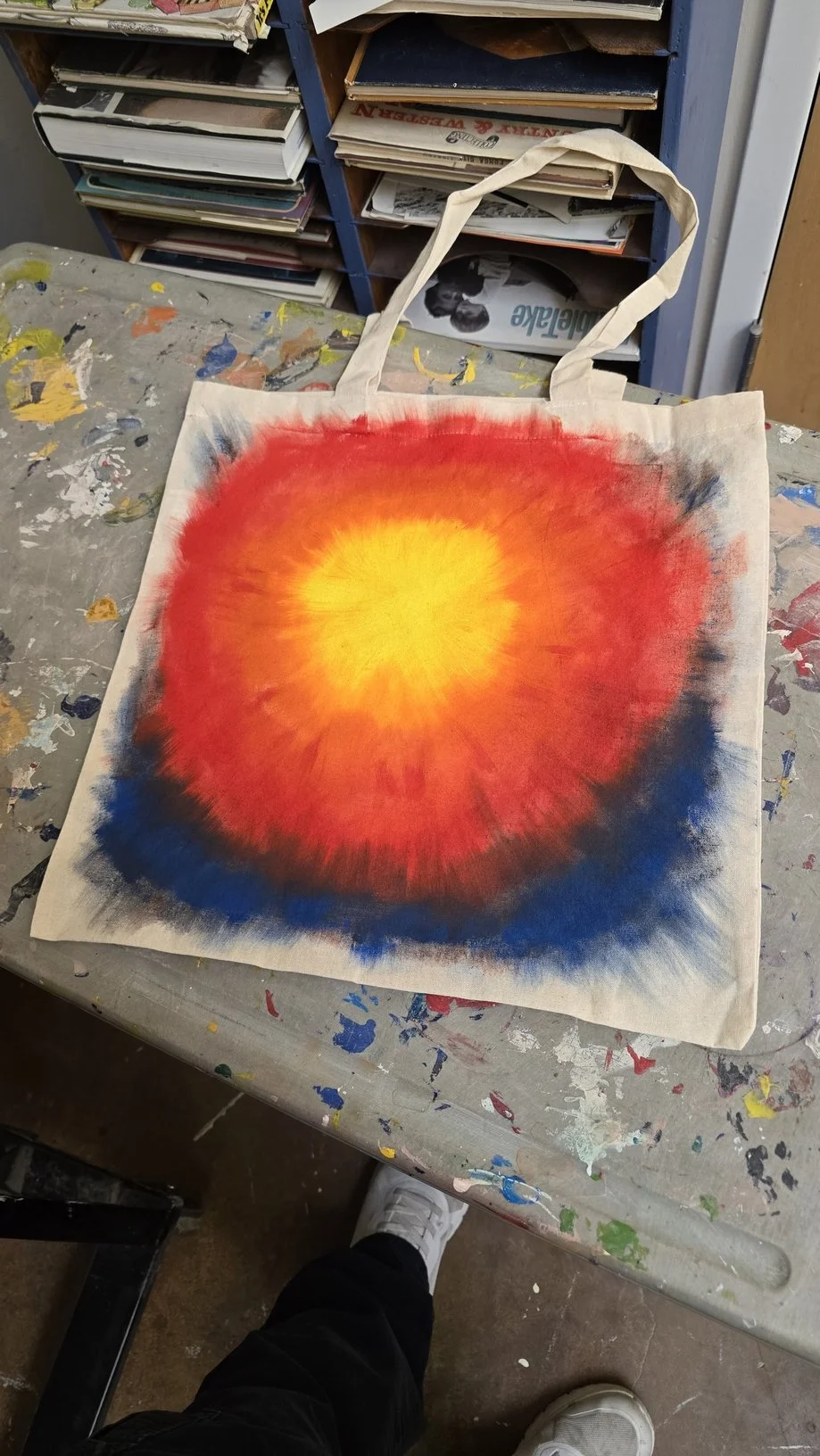 A canvas tote bag with a brightly colored burst design, featuring a yellow center, orange middle, and blue and black edges, placed on a paint-splattered table.