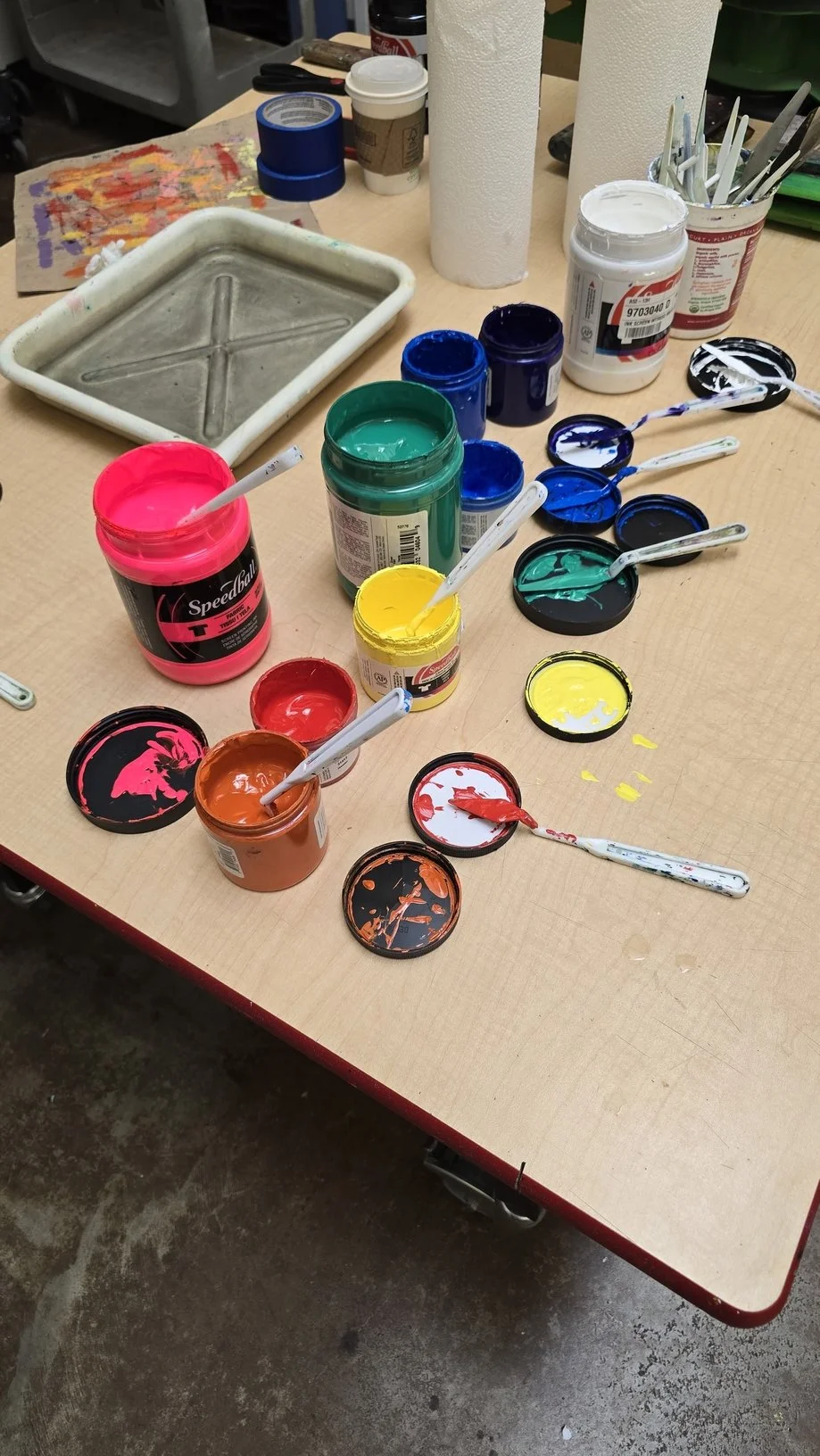 Array of open paint jars in pink, yellow, green, blue, black, orange, and white on a table with paintbrushes, paper towels, and a paint tray.