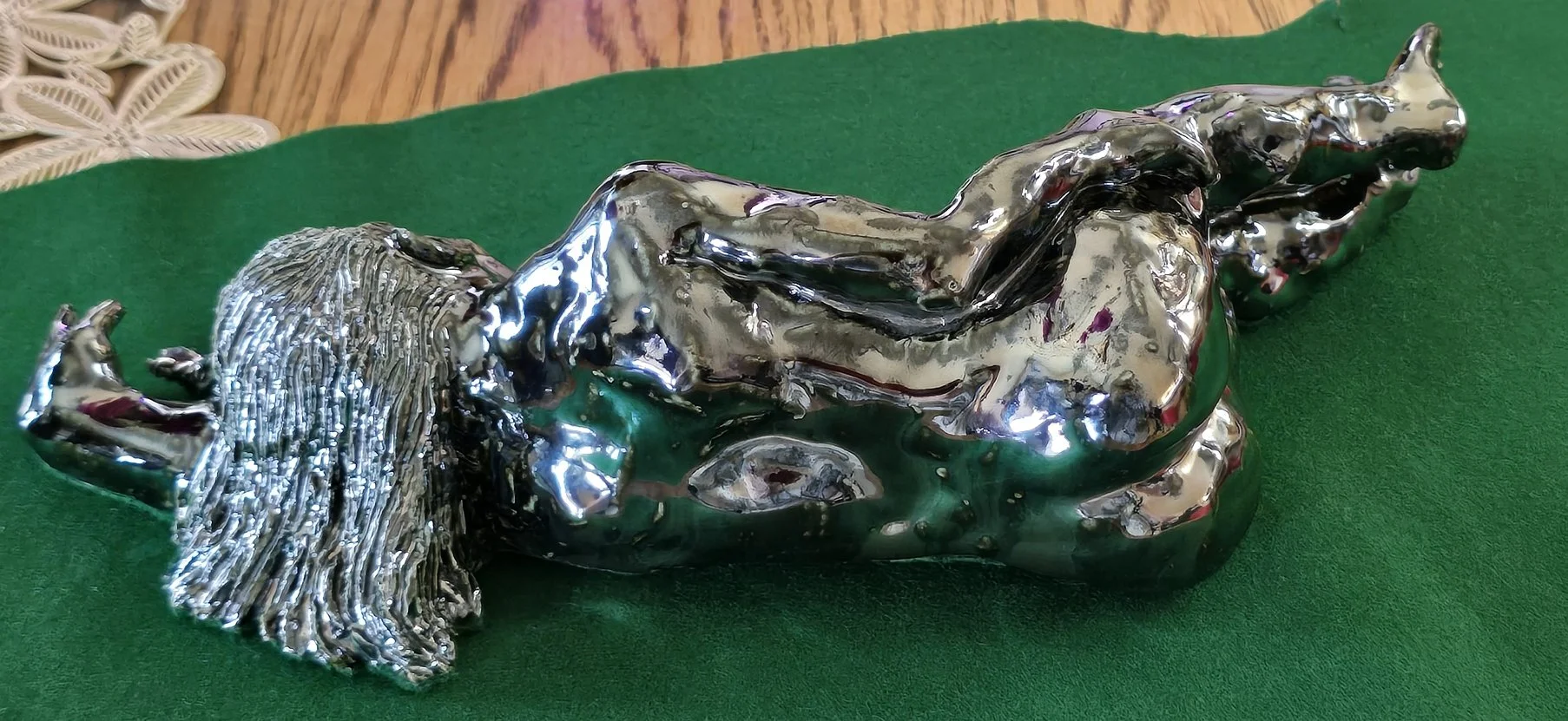 Silver- or palladium-glazed ceramic sculpture of a reclining woman with long hair, lying on her side with one arm behind her head, lying on green fabric with a wooden surface in the background.