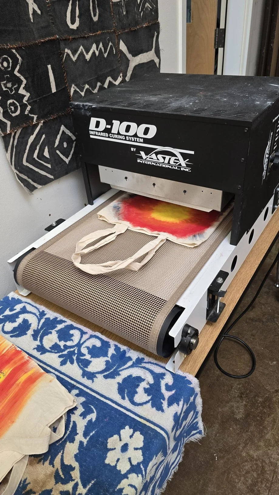 A tote bag with a tie-dye pattern in red, yellow, and black being heat pressed with a heat press machine labeled 'D-100 Infrared Curing System' by Vastek International Inc. The work surface has a blue and white floral tablecloth.