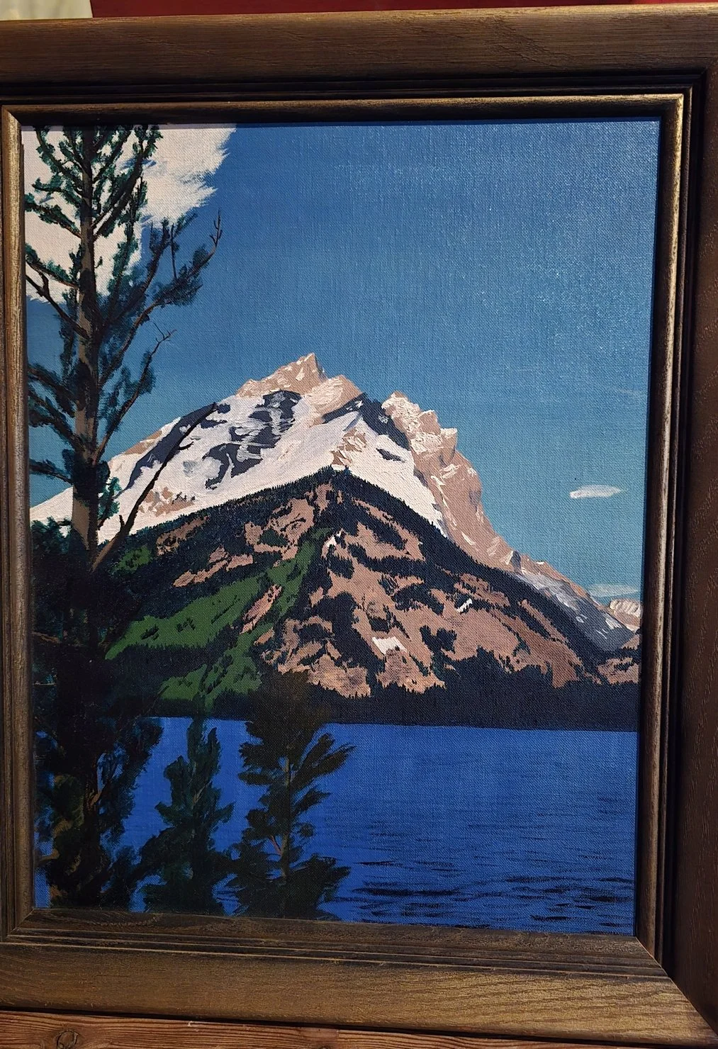 Painting of a mountain with snow-capped peak, green slopes, and a lake in the foreground, framed in dark wood.