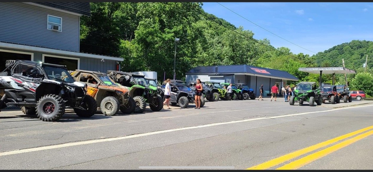 Line of off-road vehicles parked along a roadside, with people walking and socializing nearby, trees and a building in the background under a blue sky.