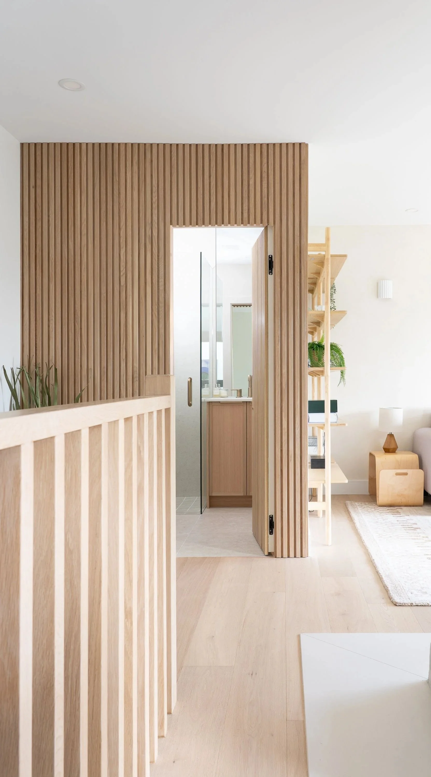 Interior view of a modern home featuring wooden slats and a sliding glass door leading to a bathroom. Light-colored wood flooring and furniture, with a cozy living space visible on the right, including a side table, lamp, and plants.