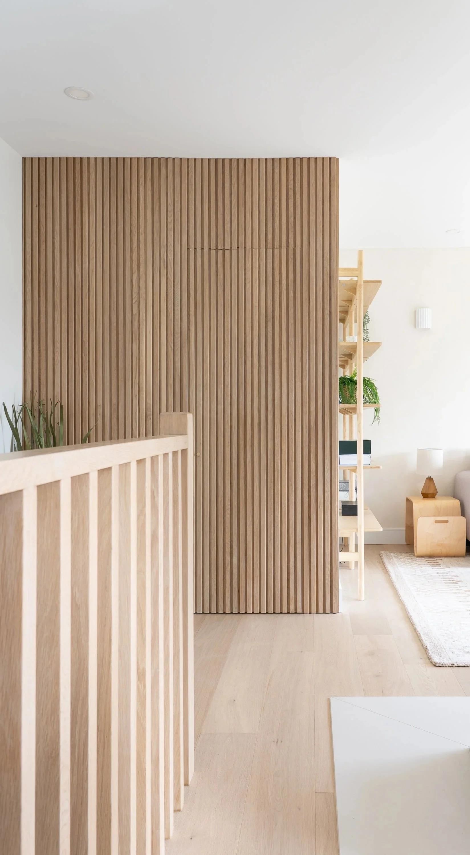 Interior of a modern home with light wood accents, including a slatted wooden partition, bookshelf with plants and books, a small side table with a lamp, and a light-colored rug on wooden flooring.
