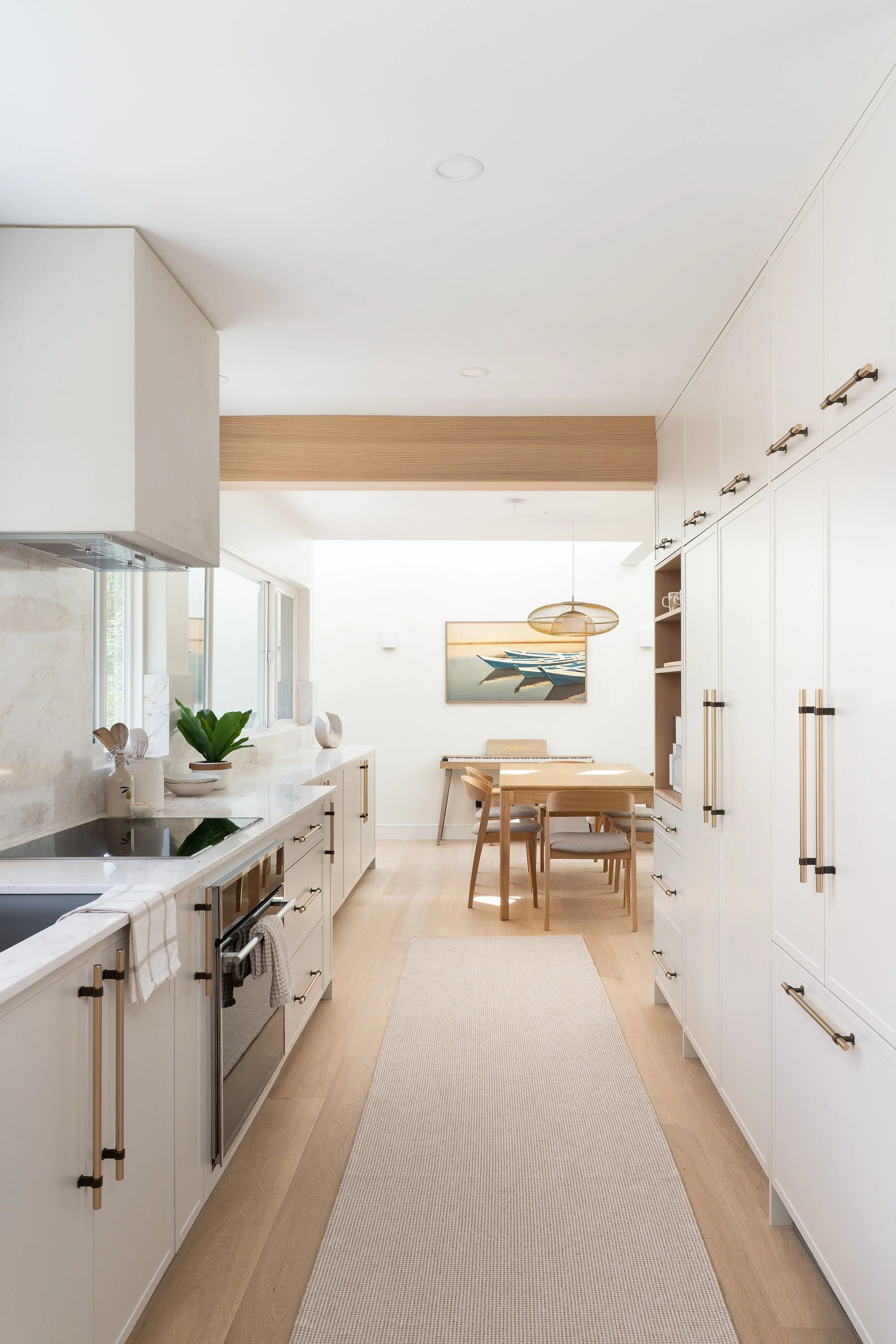 Bright, modern kitchen with white cabinets, marble countertops, and light wooden flooring. A dining area with a wooden table and chairs is visible in the background, along with artwork on the wall and a woven pendant light fixture.