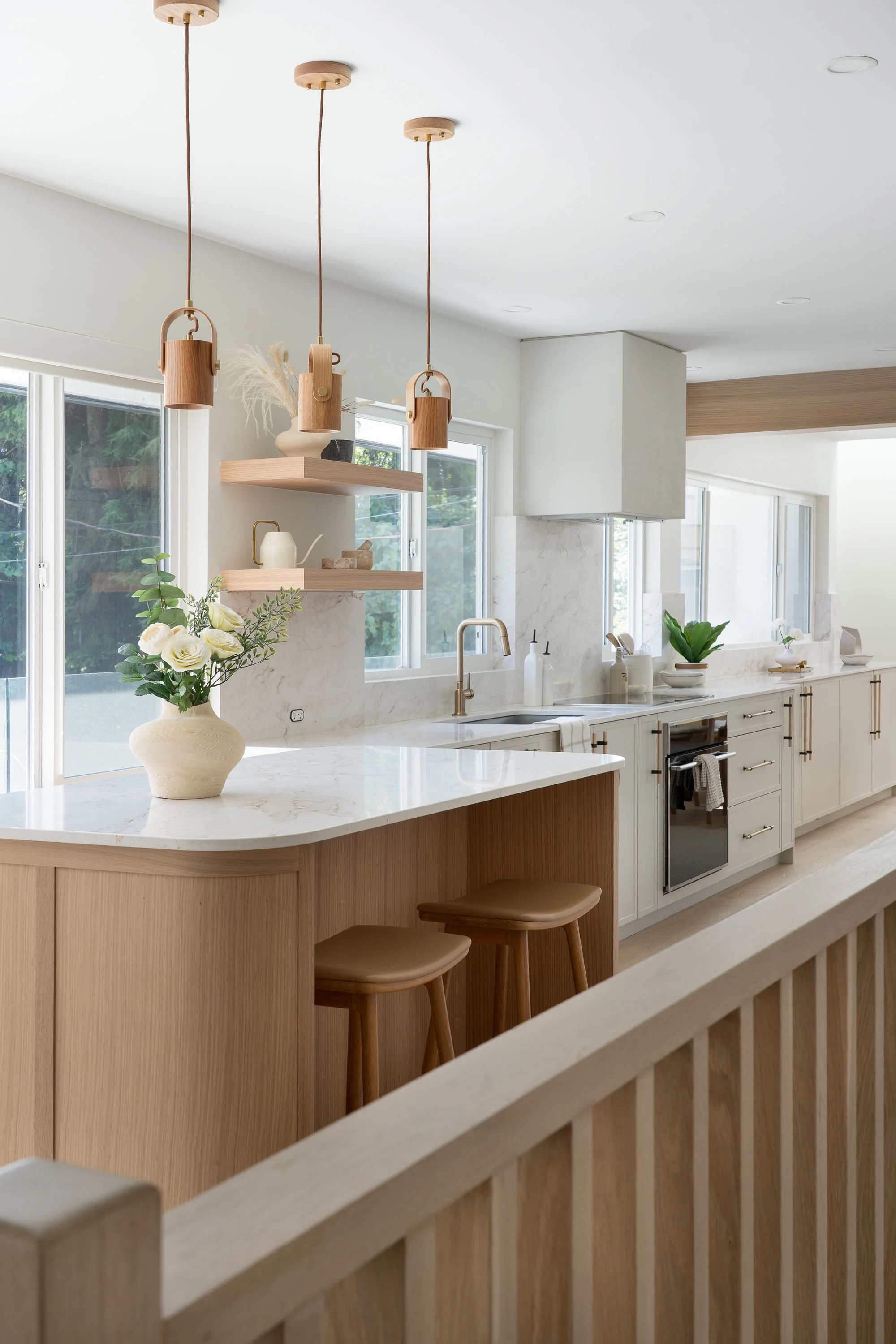 Modern kitchen with a white marble island, wooden bar stools, and hanging wooden pendant lights. There are large windows, greenery outside, and open shelves with decorative items.