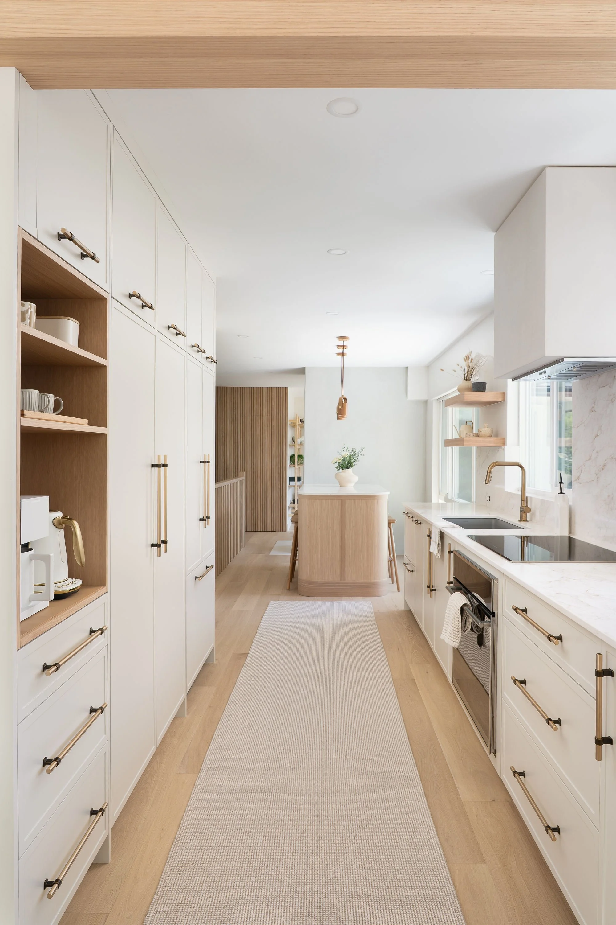 Bright, modern kitchen with white cabinets, light wood accents, and a beige runner rug on light wood flooring, illuminated by natural light from large windows.