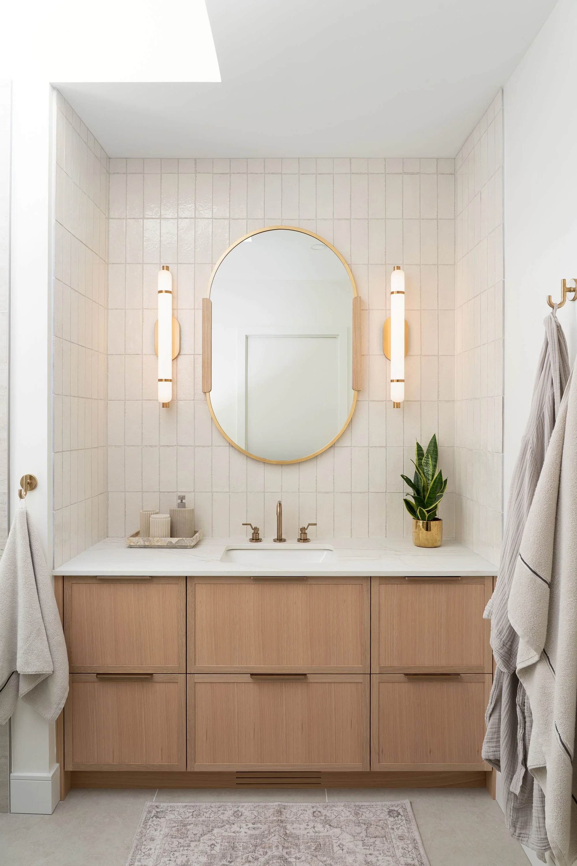 Bathroom vanity with a large oval mirror, light wood cabinet, marble countertop, and two vertically oriented wall-mounted light fixtures. There are hand towels on hooks on the sides and a potted plant on the right side of the countertop.