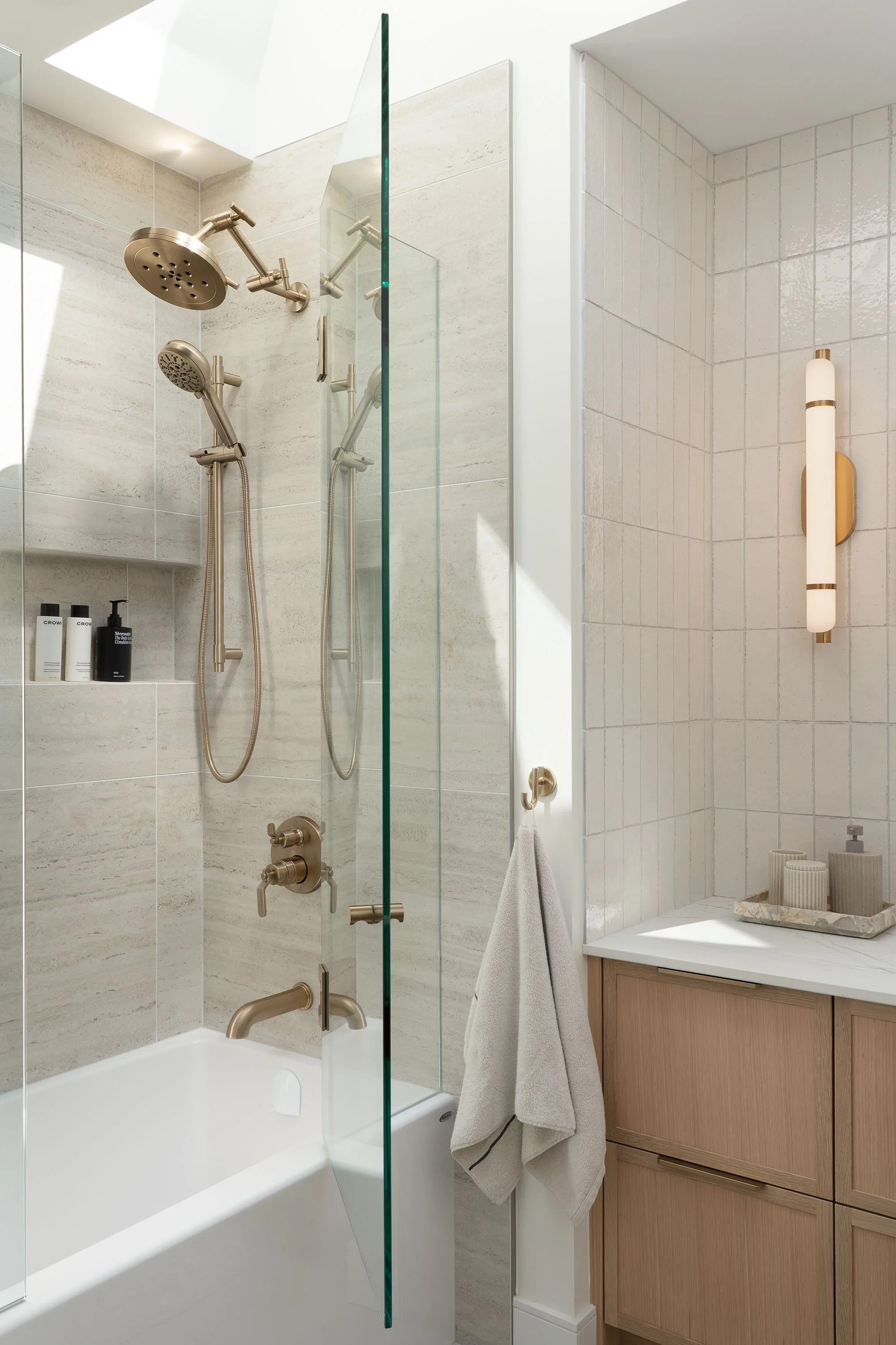 Modern bathroom with a shower area, beige tiles, a bathtub, dual showerheads, a glass divider, and a wooden cabinet with a countertop holding a decorative tray and containers.