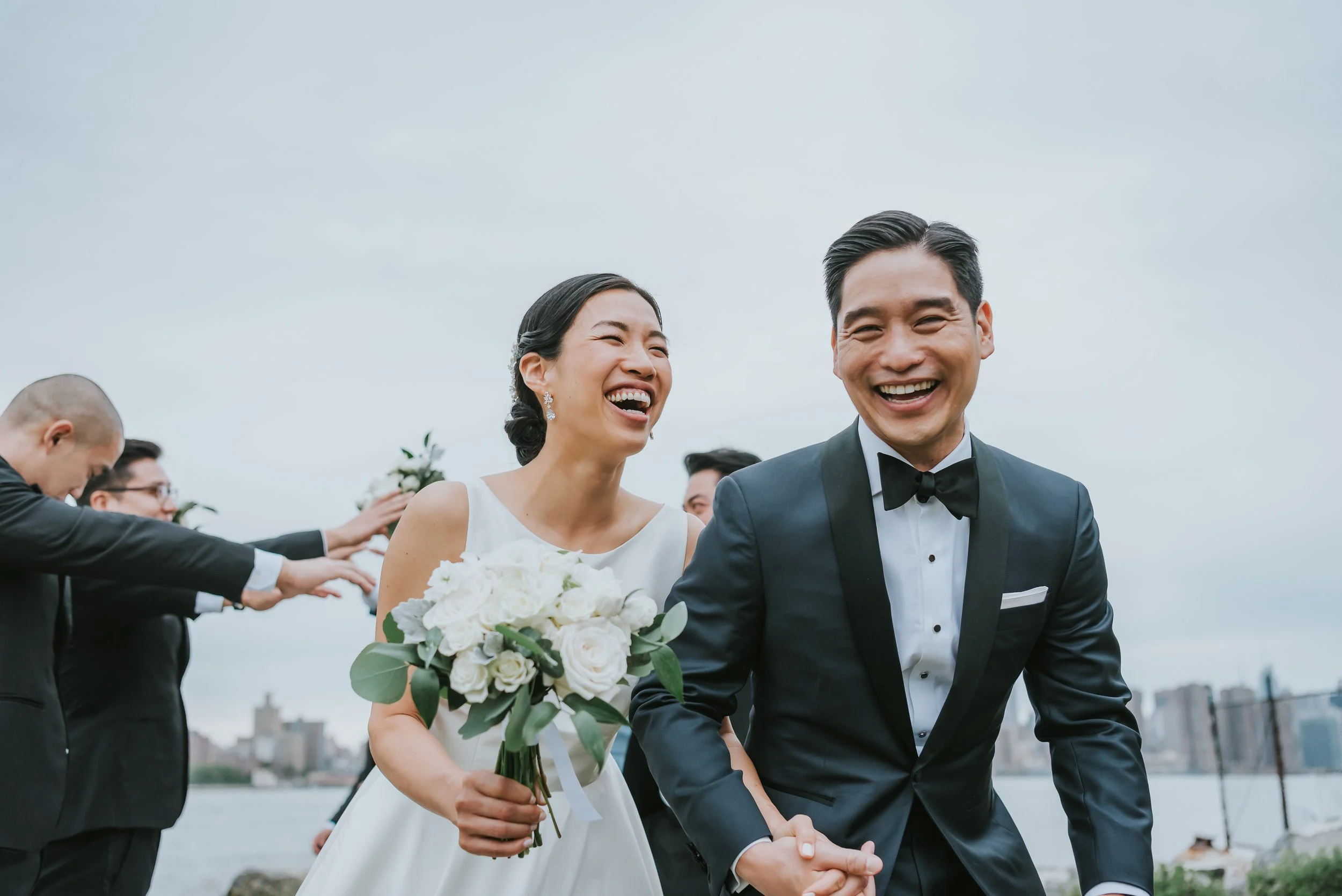 A smiling bride and groom holding hands during their wedding ceremony outdoors near a body of water, with wedding guests in the background.