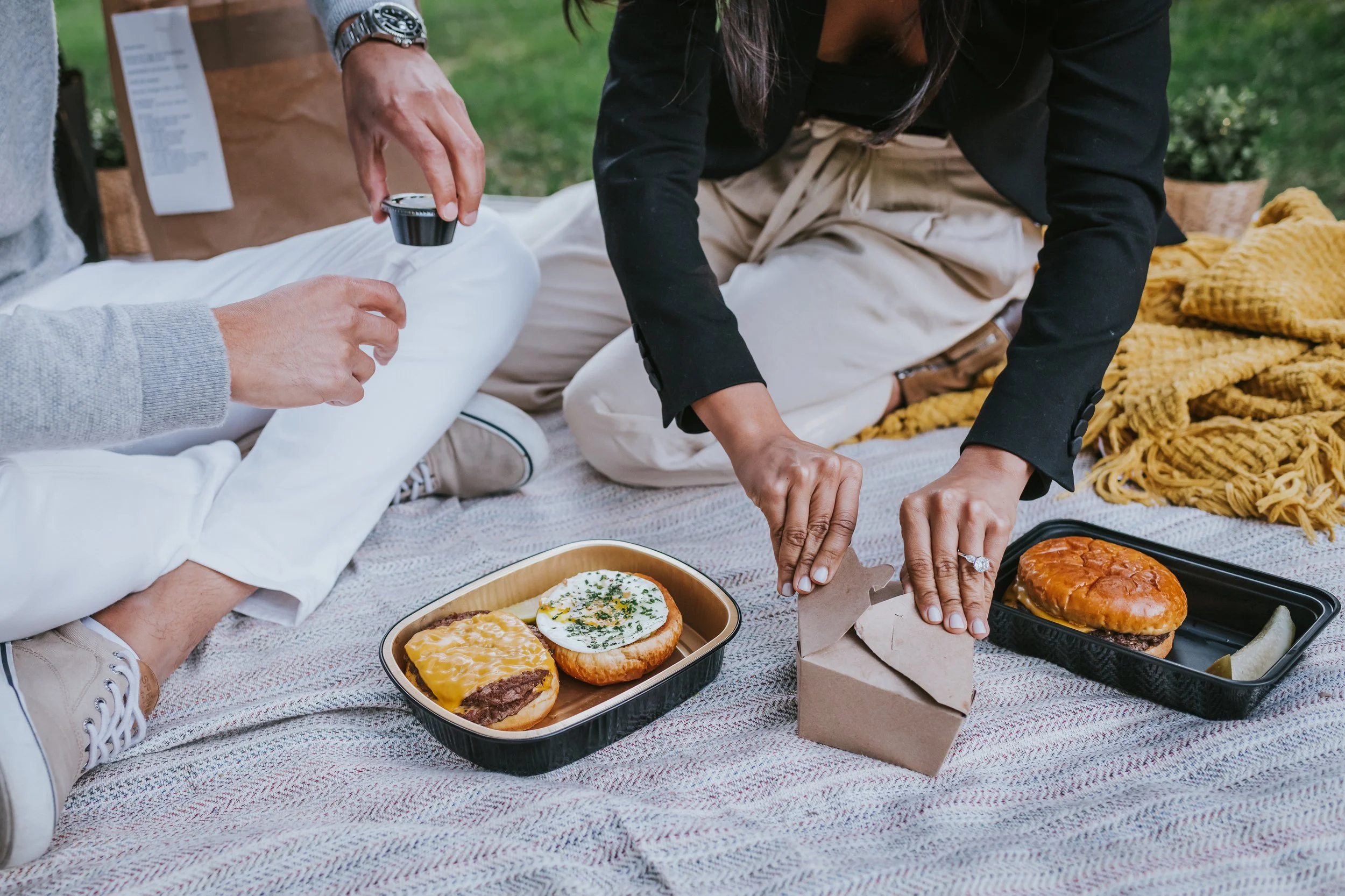 People having a picnic on a blanket outdoors, with food containers, sandwiches, and picnic items, some person is handling a takeout box.