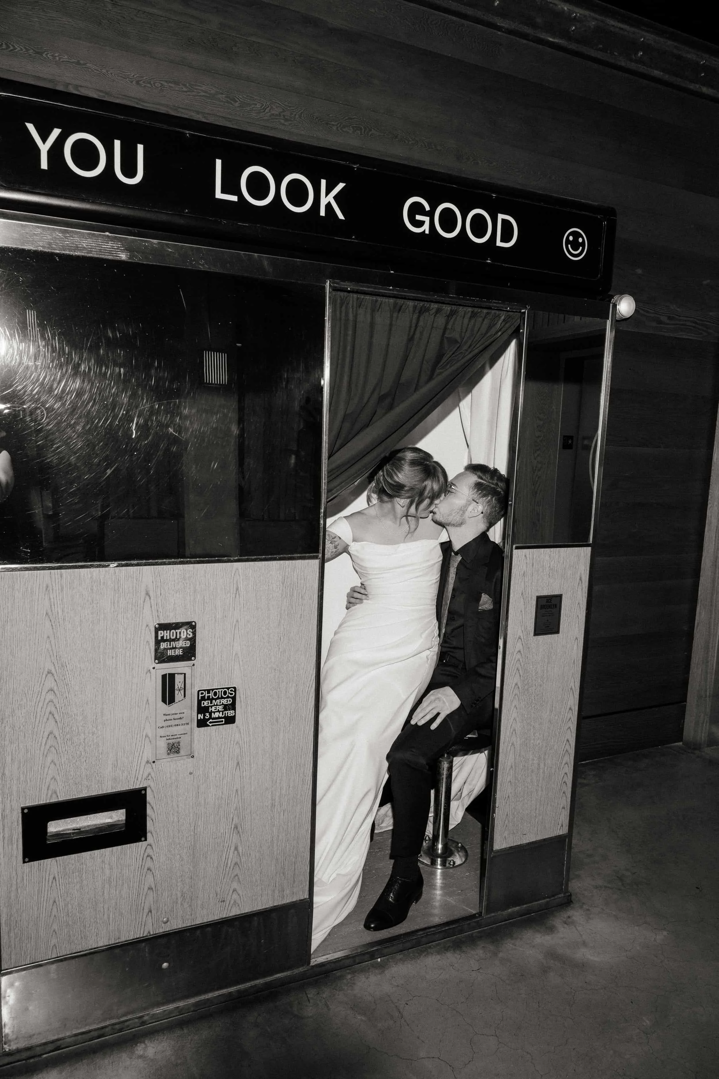 A black and white photo of a photo booth with a sign saying 'You Look Good' and a smiley face. Inside, a bride and groom are kissing; the bride wears a white wedding dress, and the groom is in a black suit. They are sitting close, sharing a kiss.