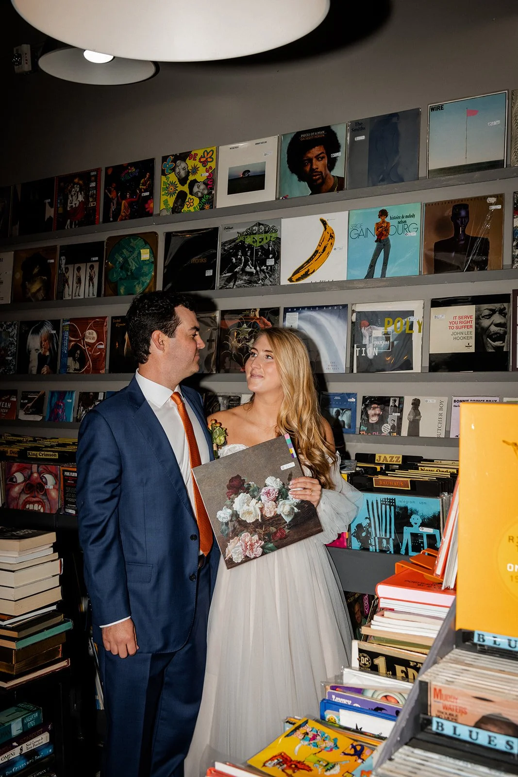 A man in a blue suit and a woman in a white dress are inside a record store, standing close and looking at each other. The woman is holding a painting of flowers. The store has shelves filled with vinyl records and books.