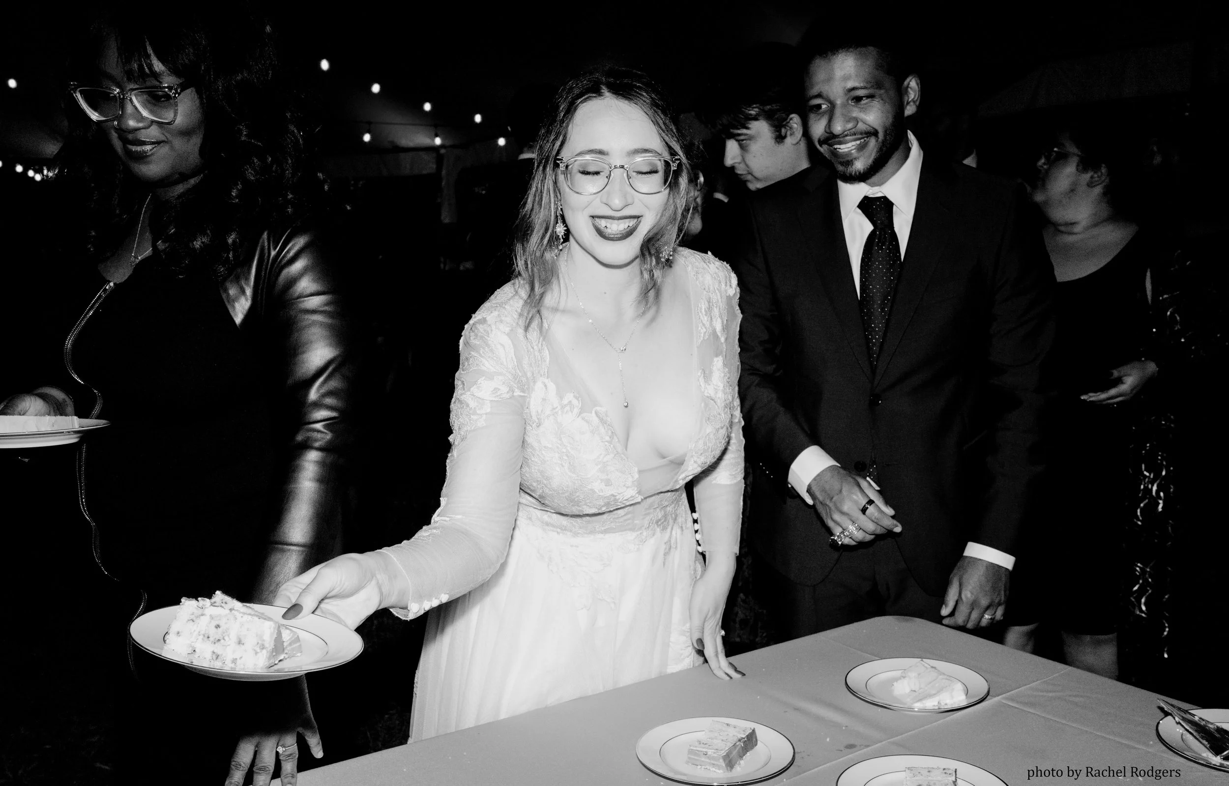 A woman in a wedding dress smiling and holding a plate of cake next to a man in a suit at a celebration, with other guests in the background.