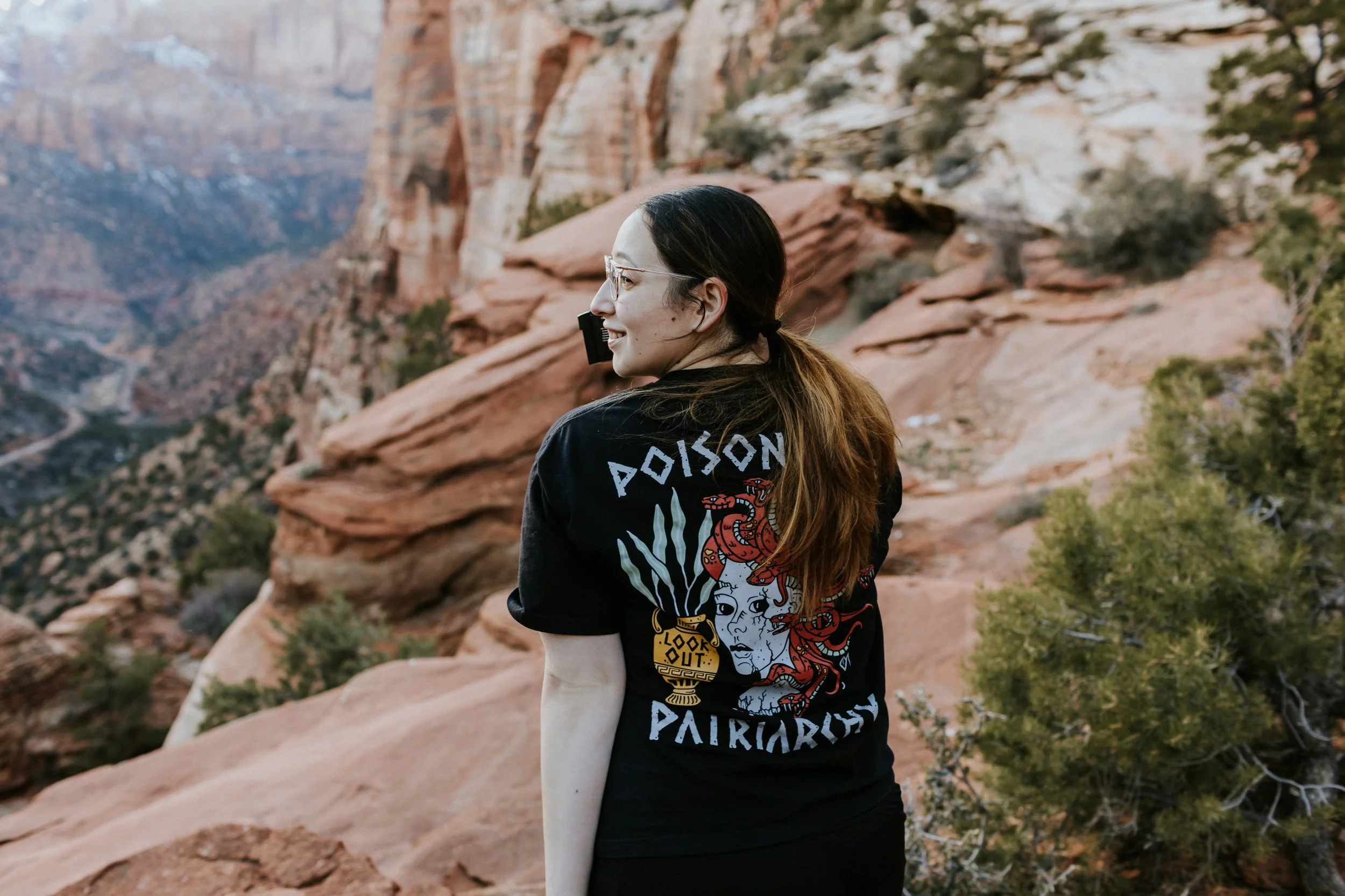A woman with glasses and long brown hair tied in a ponytail standing outdoors in a desert landscape with red rocks and sparse green bushes, smiling and looking to her left, wearing a black t-shirt with a graphic and the words "Poison Patriot" on the back.