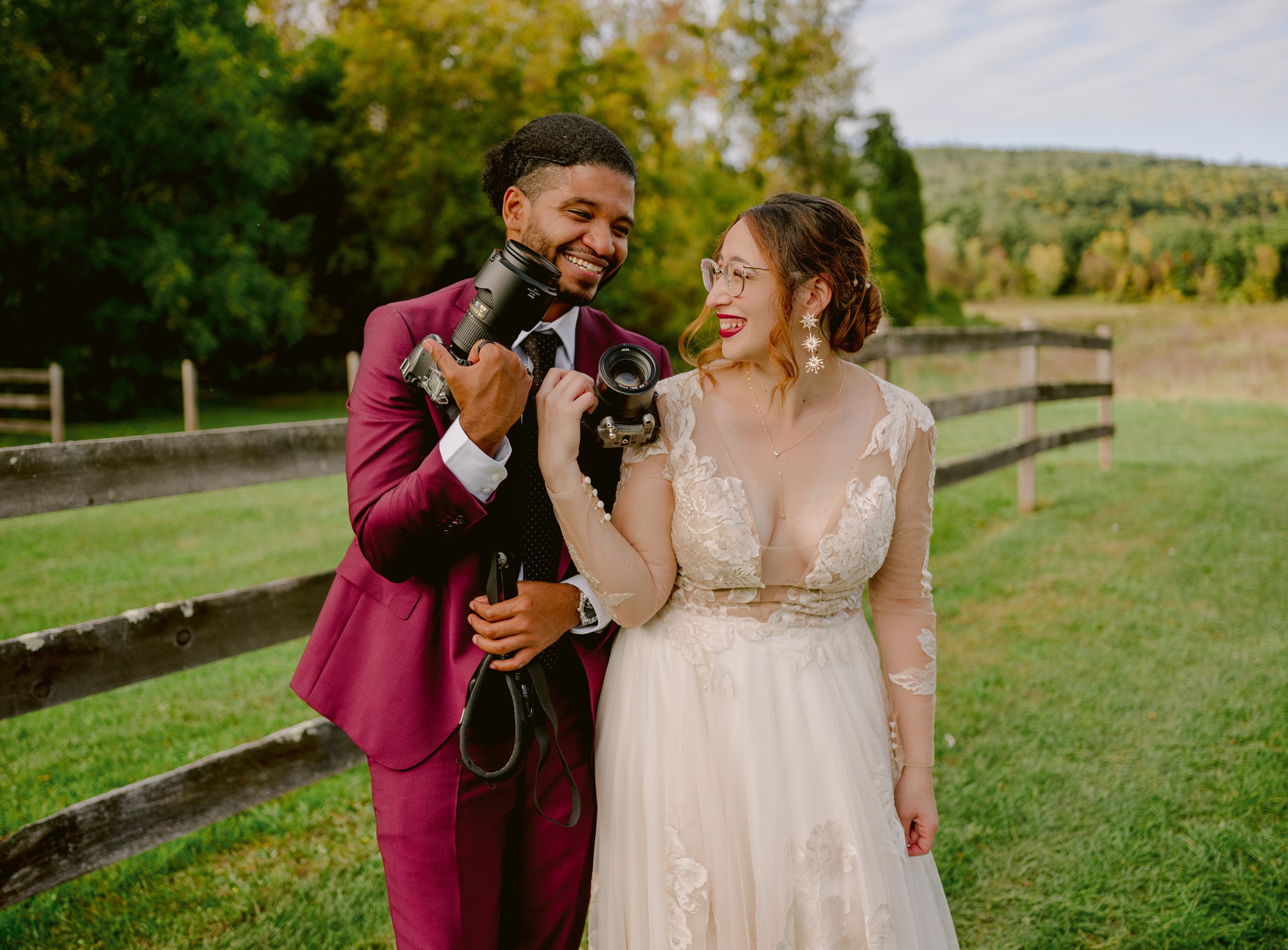 A smiling man and woman in wedding attire standing outdoors near a wooden fence, looking at each other with a camera in hand, surrounded by green grass and trees with autumn foliage.