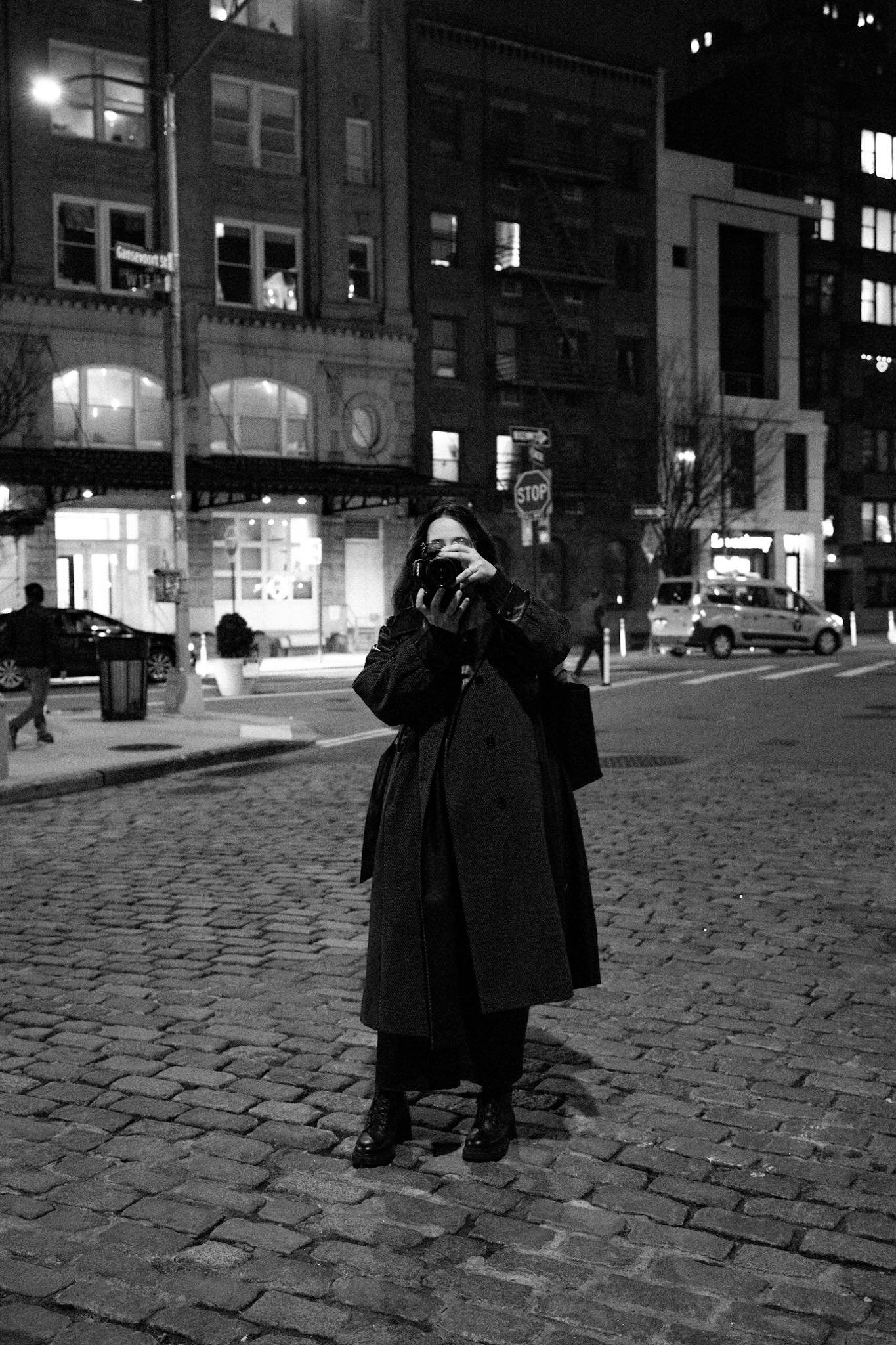 A person dressed in dark clothing, including a long coat and boots, stands on a cobblestone street at night, taking a photo with a camera. The background shows urban buildings, streetlights, parked cars, and a stop sign, all in black and white.