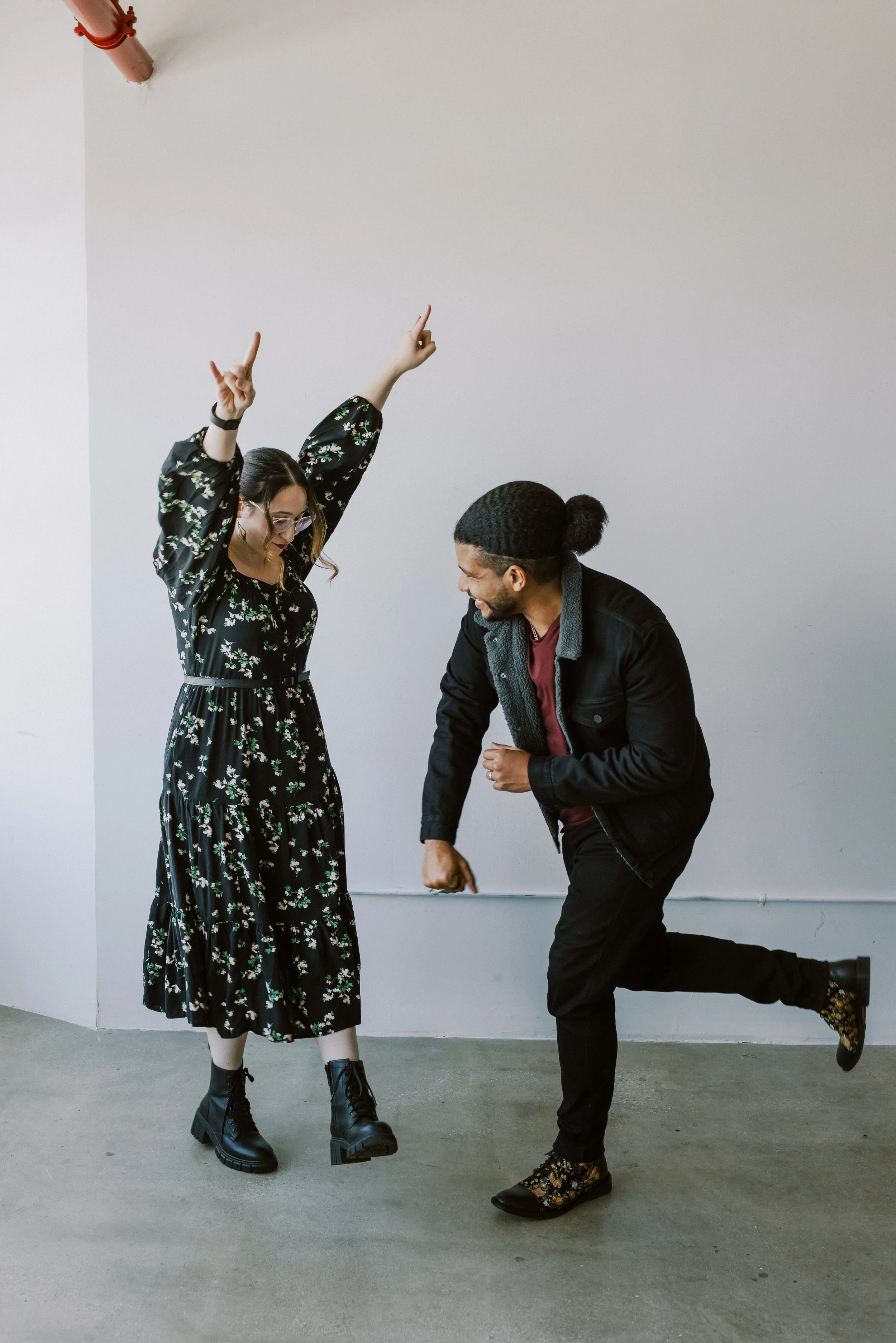 A woman with glasses in a floral dress and black boots dancing with a man with a bun and black boots with floral pattern, both smiling and energetic in a plain indoor space.