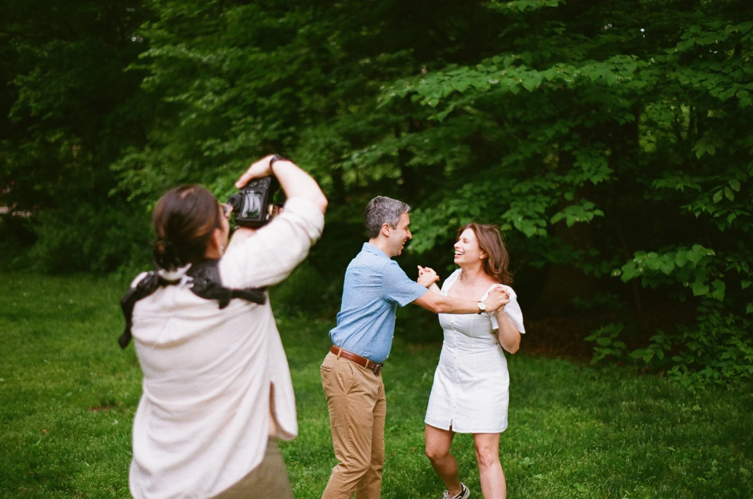 A man and woman joyfully dancing outdoors on grass with greenery in the background while a photographer captures the moment
