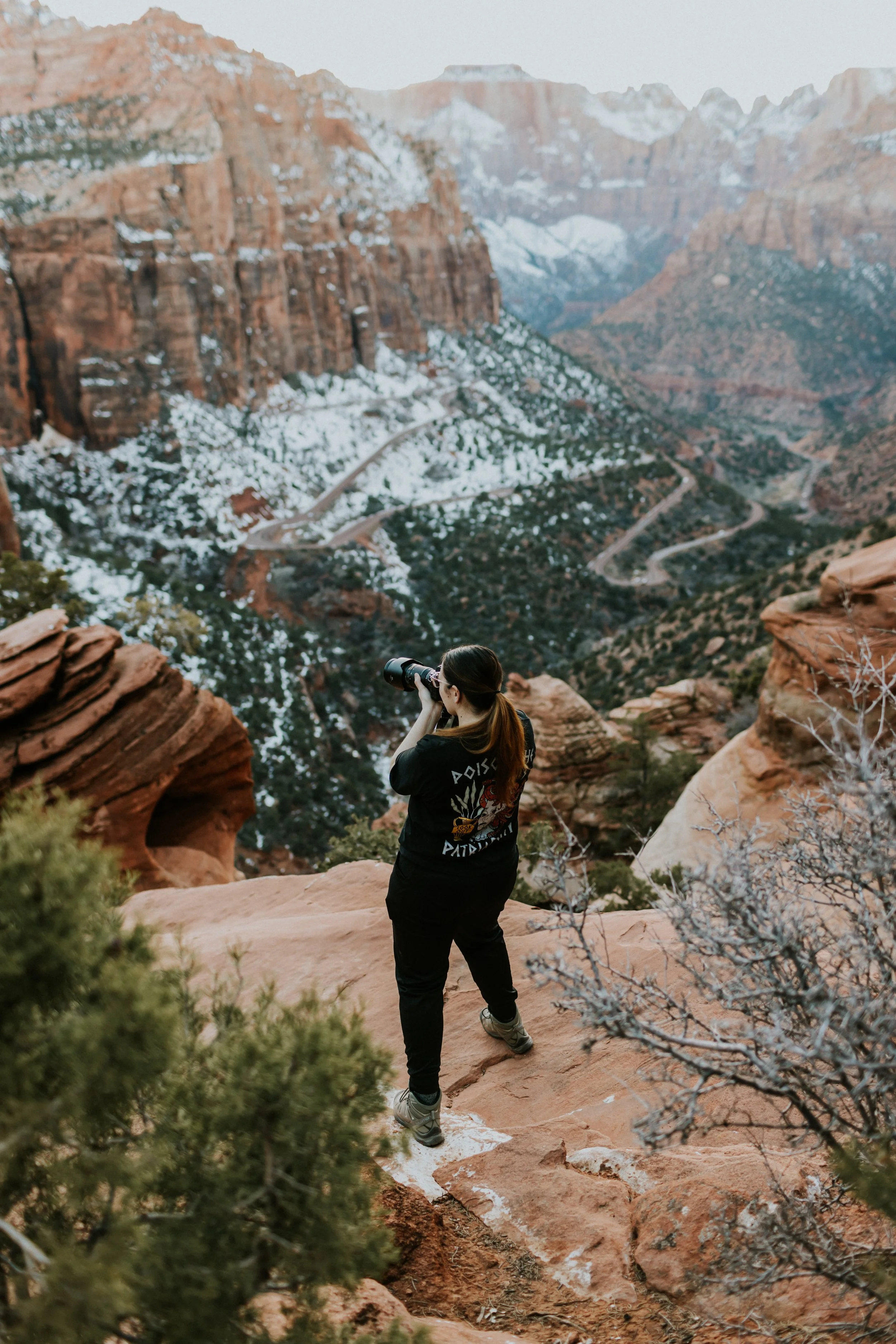 A woman with a camera taking photos of a canyon landscape with snow-covered cliffs and a winding road.