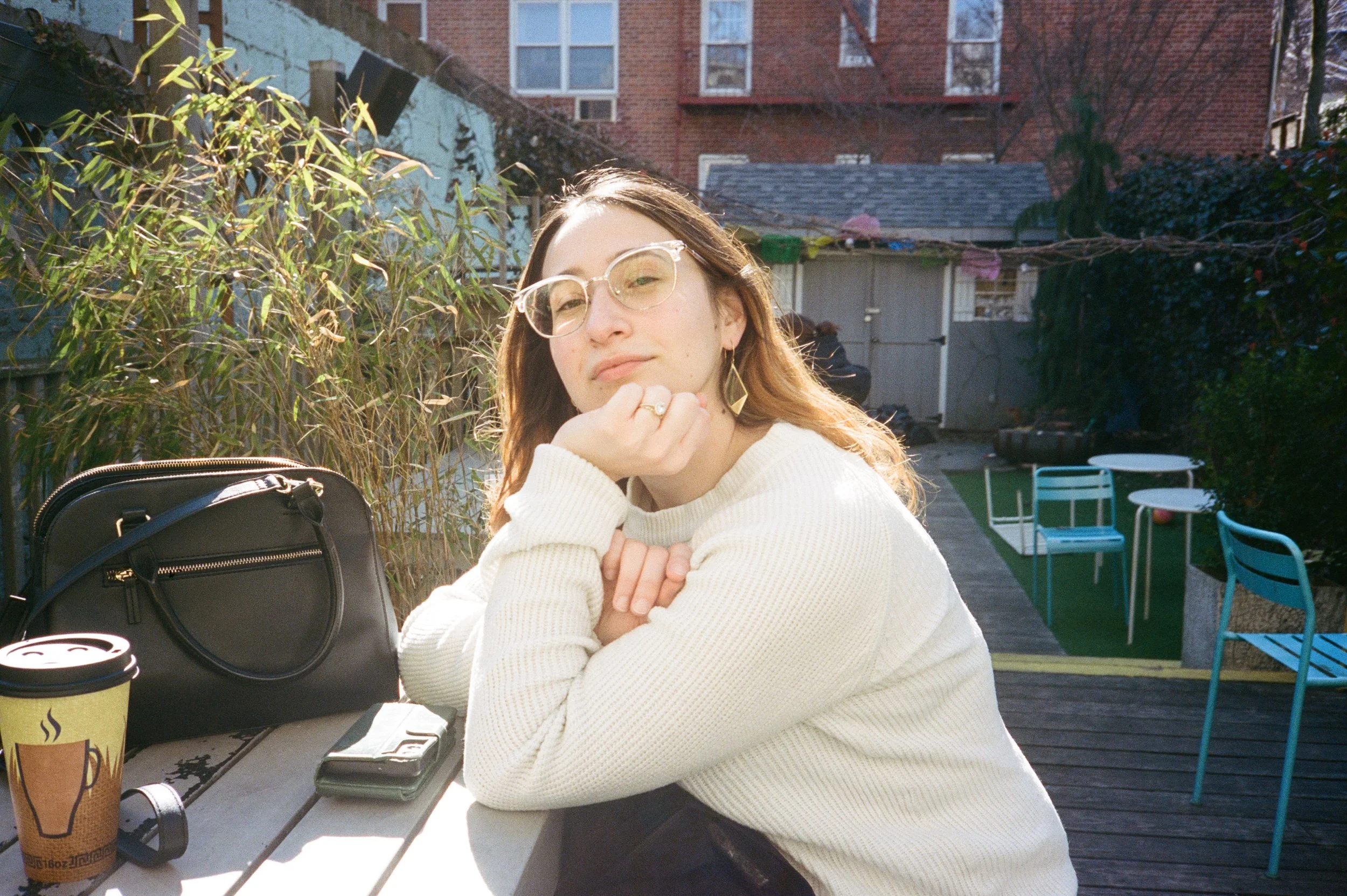 A young woman with long brown hair, wearing glasses and a cream sweater, sitting at an outdoor table on a patio with a black purse, a paper coffee cup, and a phone in front of her. She rests her chin on her hand, gazing at the camera, with sunlight i