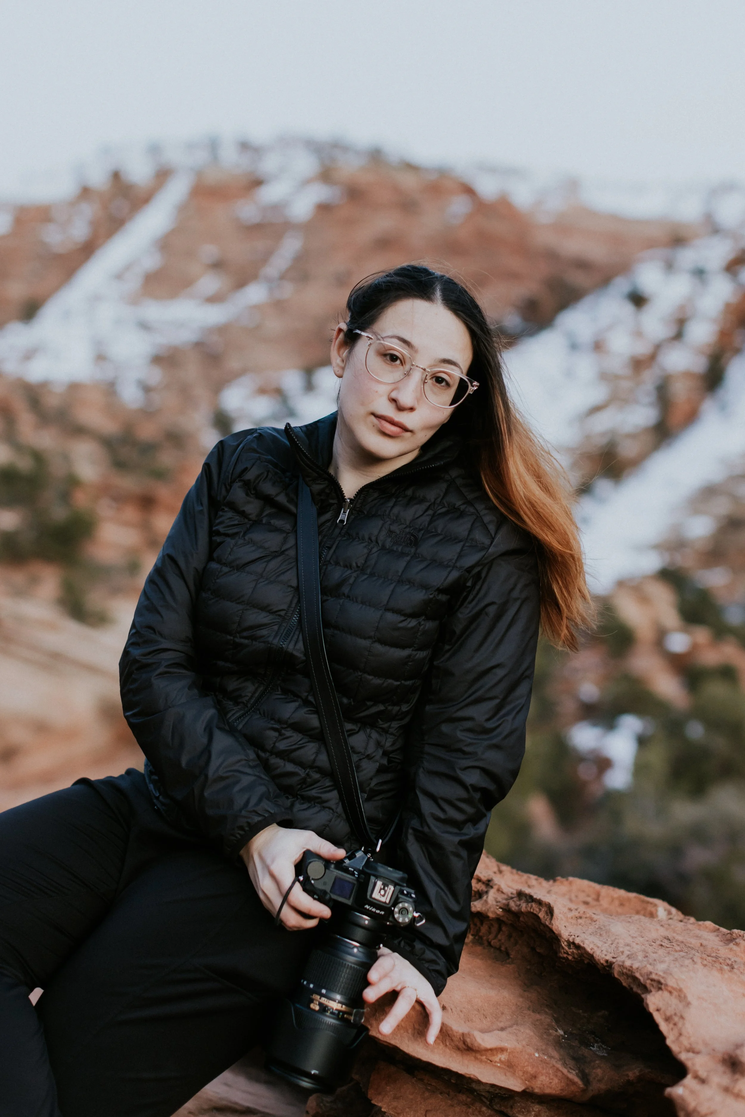 A woman with glasses and long hair sitting outdoors in a rocky landscape with snow patches, holding a camera.