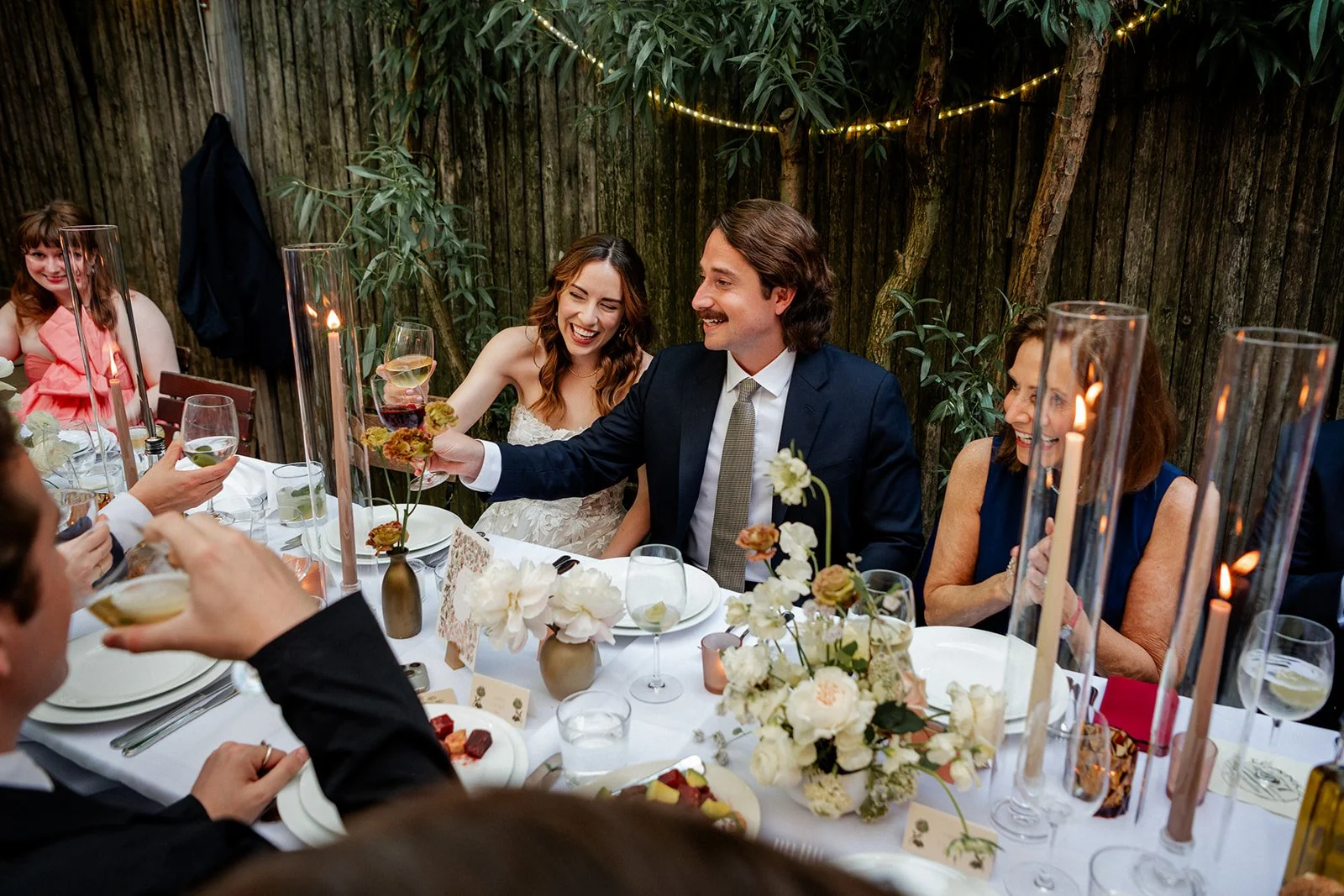 People celebrating around a dining table with flowers and candles, toasting with wine glasses at a festive outdoor party.