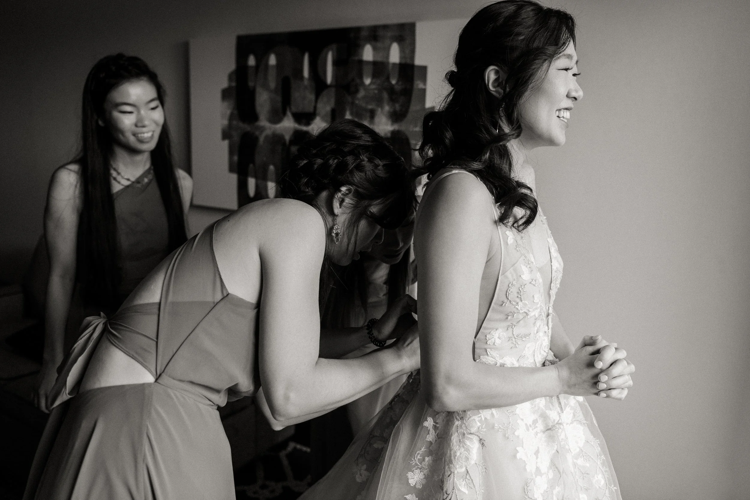A bride in a lace wedding dress smiling with her hands clasped, while three women, dressed in elegant attire, assist her with her dress. One woman leans in to adjust the dress, another smiles in the background, and the third is partially visible.