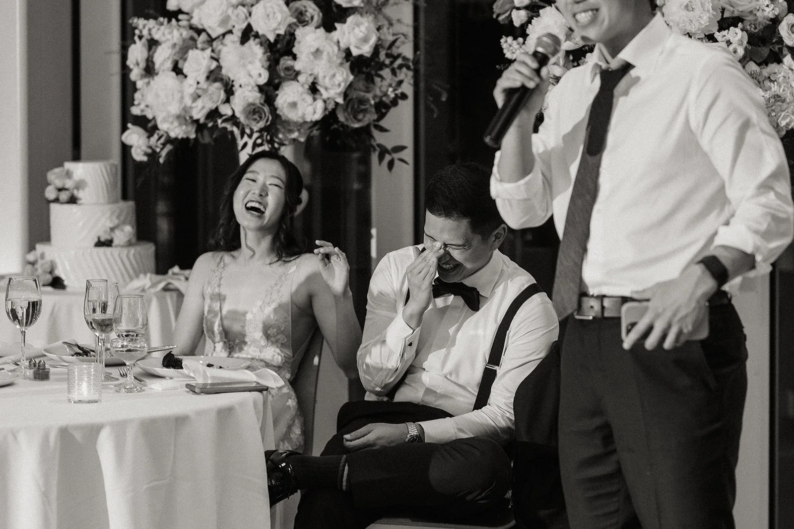 People laughing and smiling at a wedding reception, with a woman in a dress, and two men in dress shirts, one holding a microphone and the other wiping his eye, at a table with glasses and flowers.