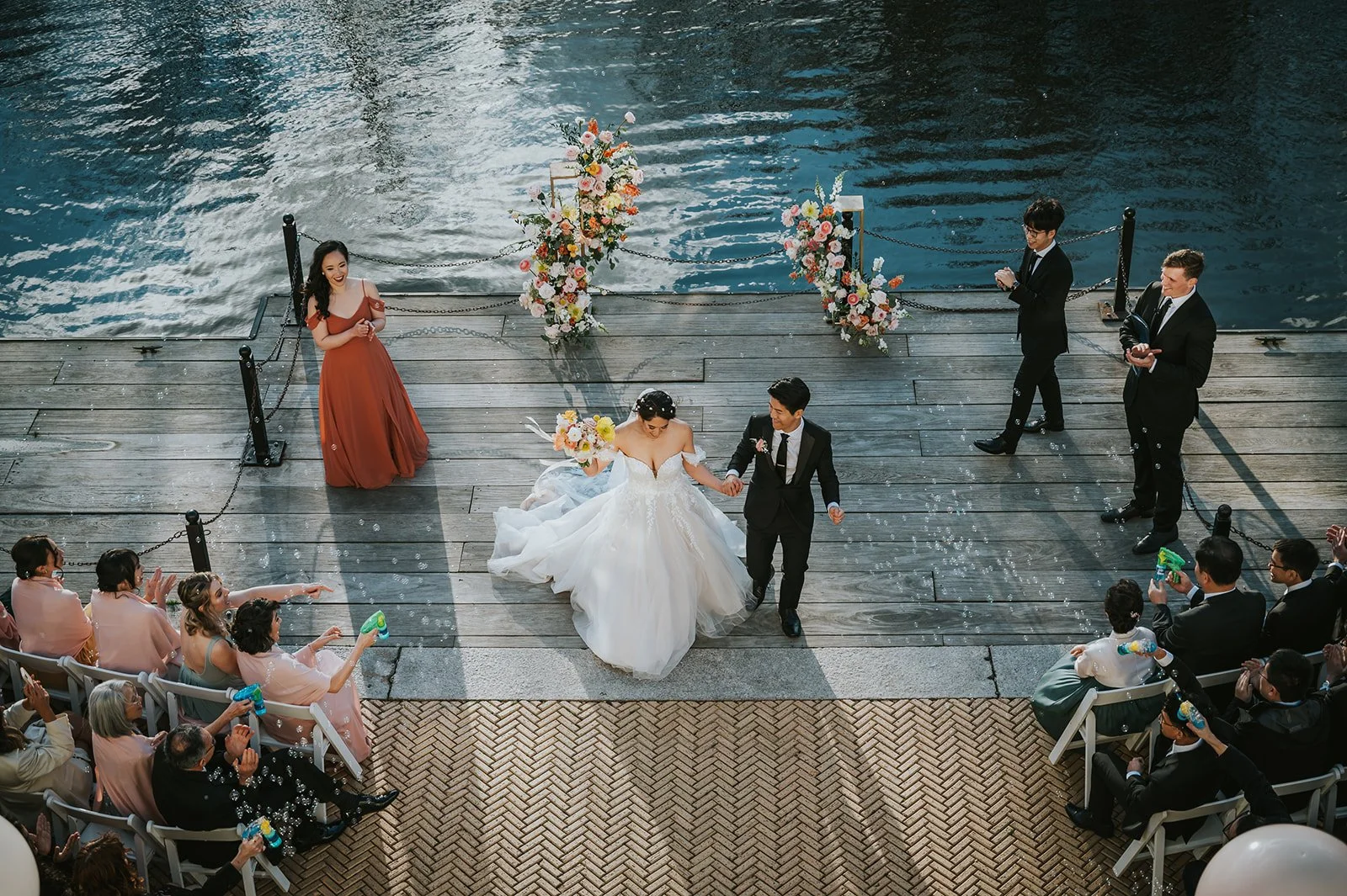 A bride and groom dancing on a wooden dock by the water during their wedding ceremony, surrounded by guests and floral decorations.