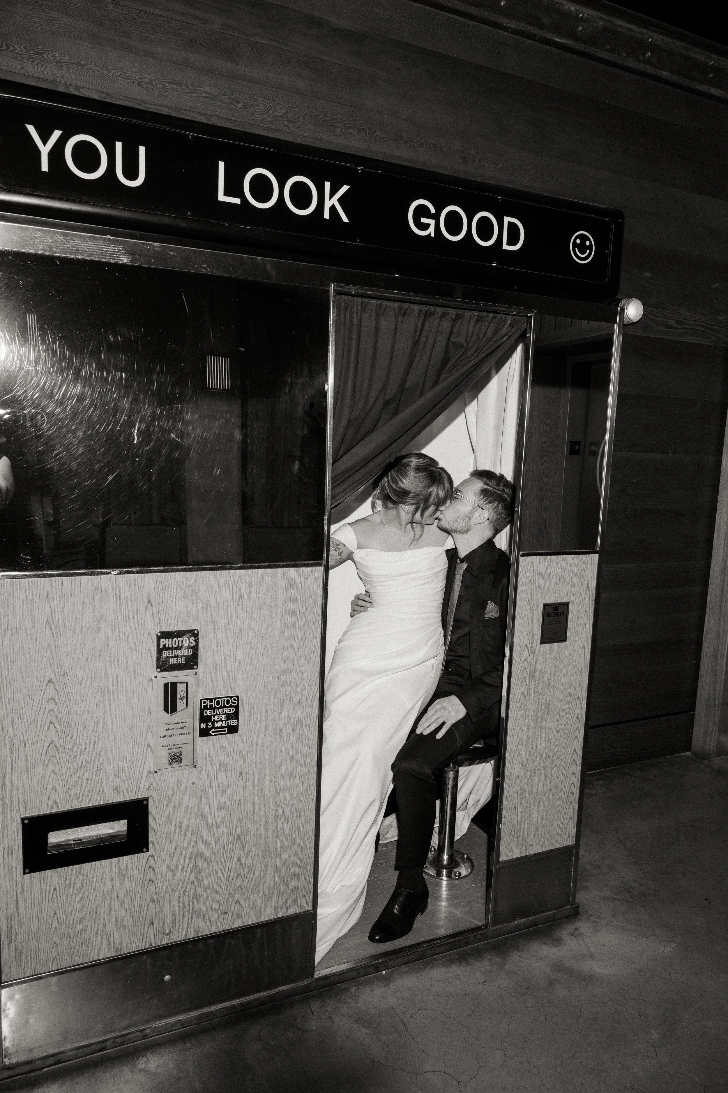 A couple in wedding attire kissing in a photo booth, with the woman in a white dress and the man in a dark suit, above a sign that says 'You Look Good' with a smiley face.