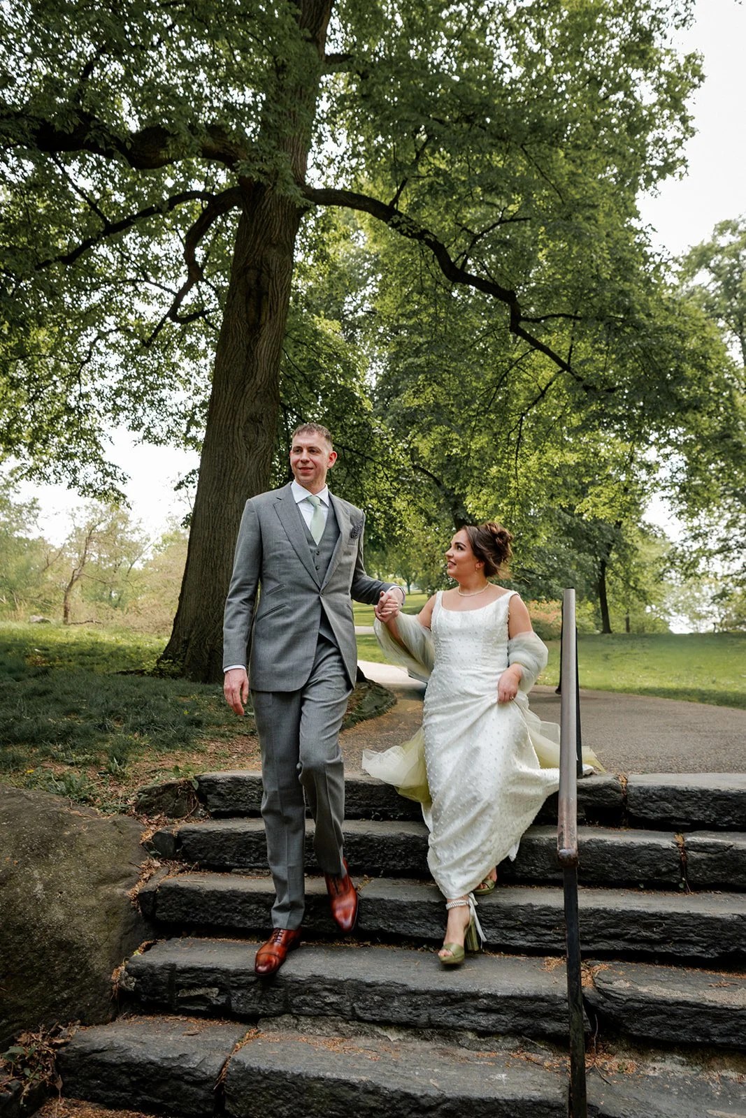 A bride and groom walking down stone steps outdoors in a park, holding hands, surrounded by green trees.