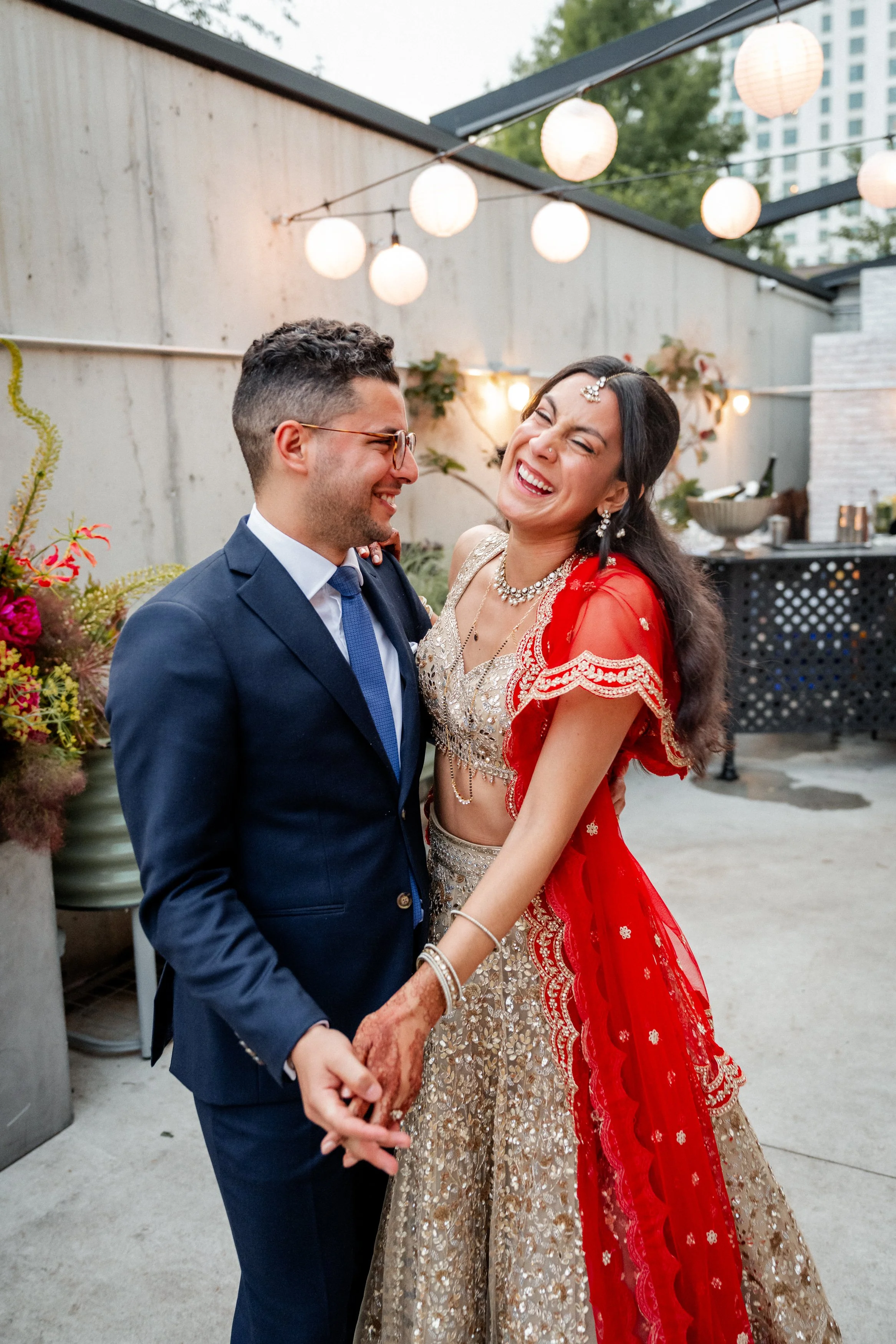 A couple dressed in traditional Indian attire dancing and laughing outdoors in a decorated venue with hanging lanterns and flowers.