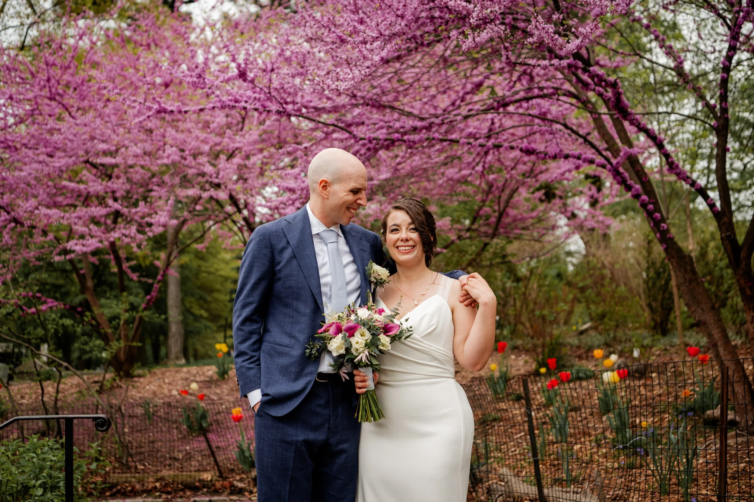 A bride and groom standing together outdoors with pink blooming trees in the background. The groom is wearing a blue suit and the bride is in a white wedding dress holding a bouquet of flowers. They are smiling and looking at each other.