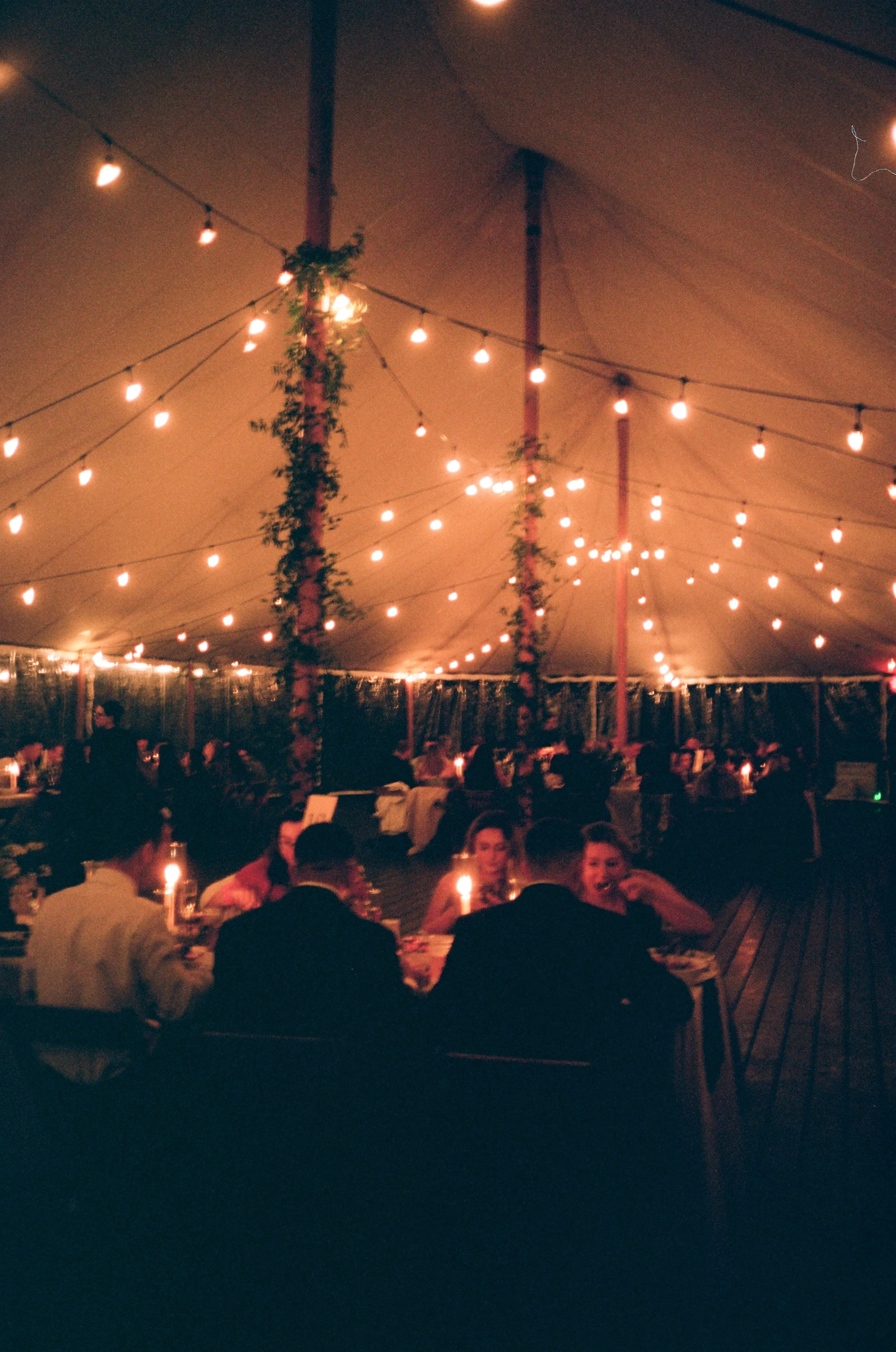 People dining at a candlelit event under a large tent decorated with string lights and hanging greenery.