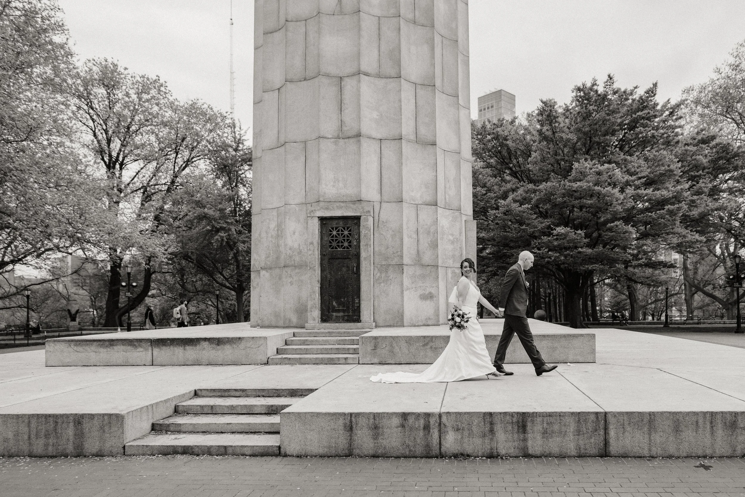 A bride in a white wedding dress holding a bouquet and a groom in a suit walk hand in hand in an urban park near a large monument, with trees and a few park visitors in the background.