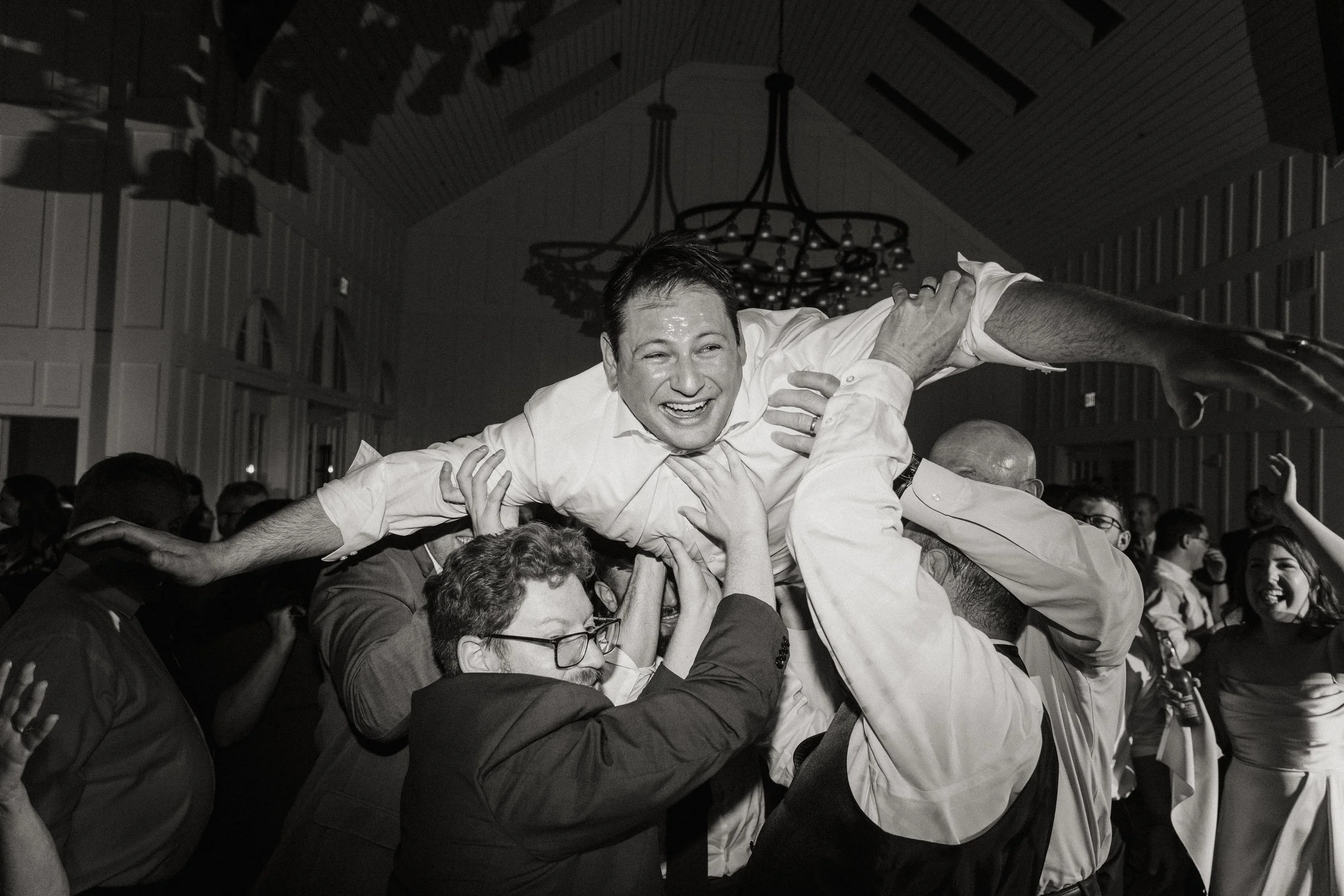 People celebrating a joyous event indoors, lifting a smiling man in a white shirt above their heads.