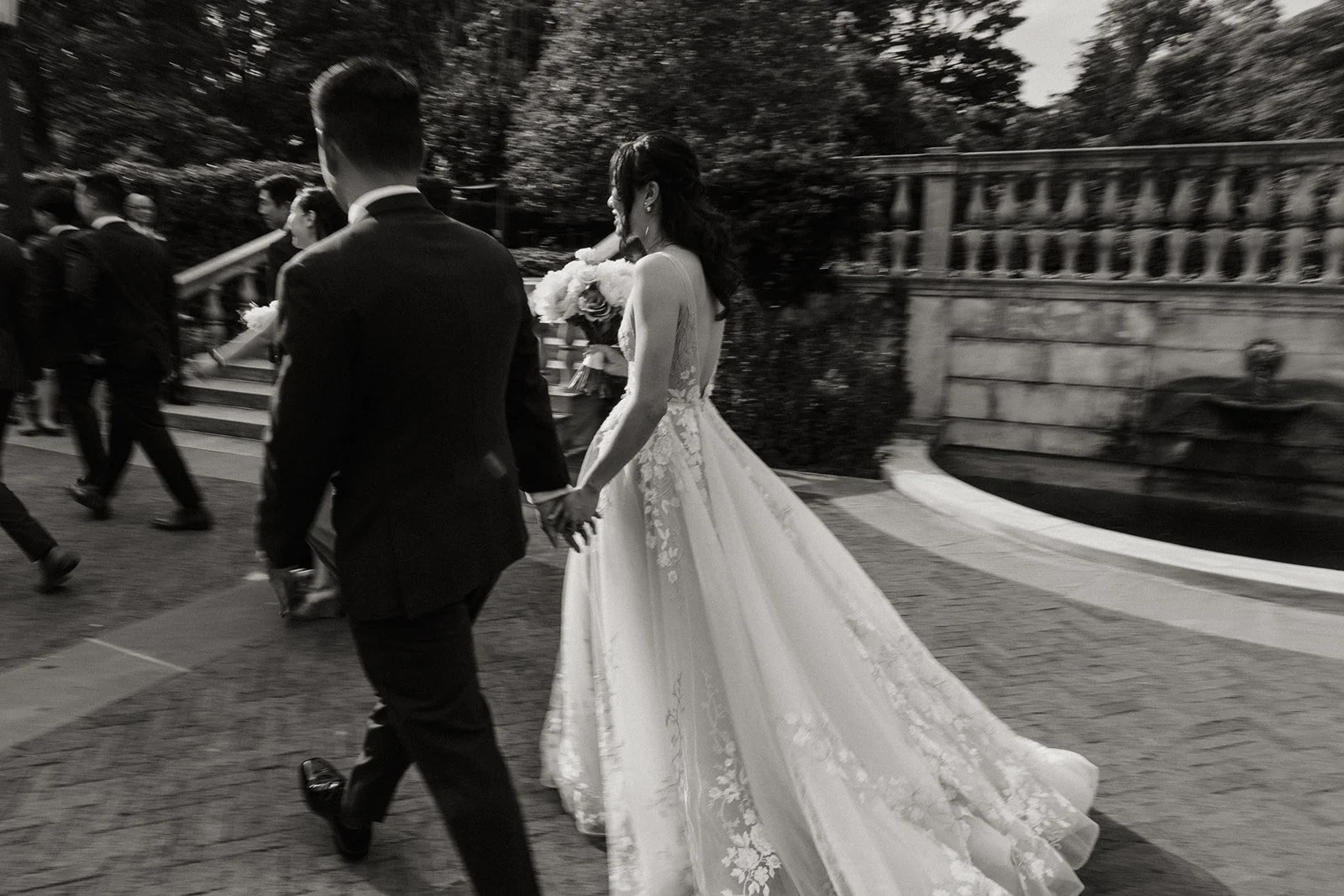 A bride and groom holding hands walk together outdoors, surrounded by other people, during a wedding ceremony at dusk or evening.