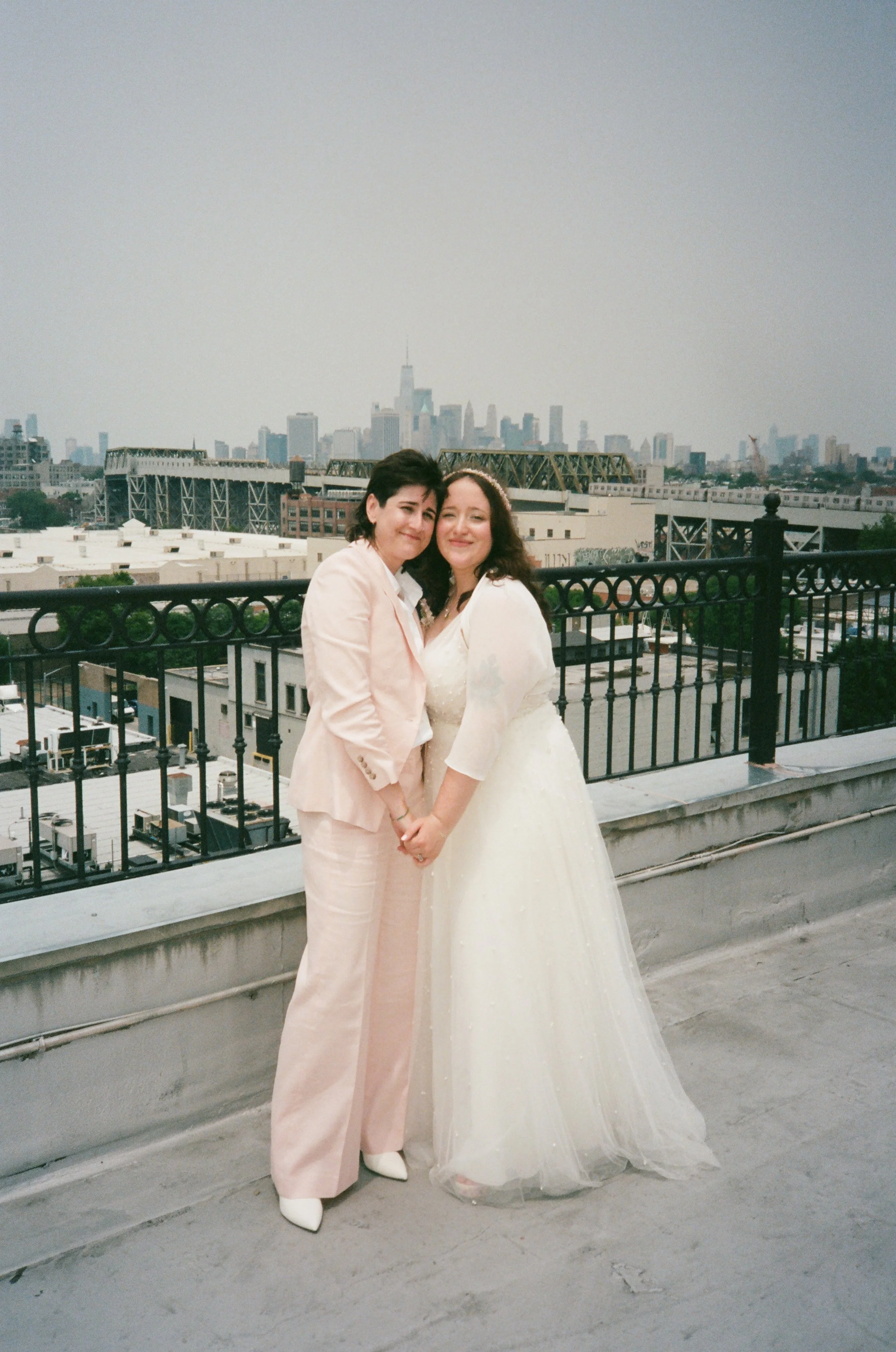 Two women holding hands and smiling on a rooftop with the New York City skyline in the background, one in a white wedding dress and the other in a light pink suit.