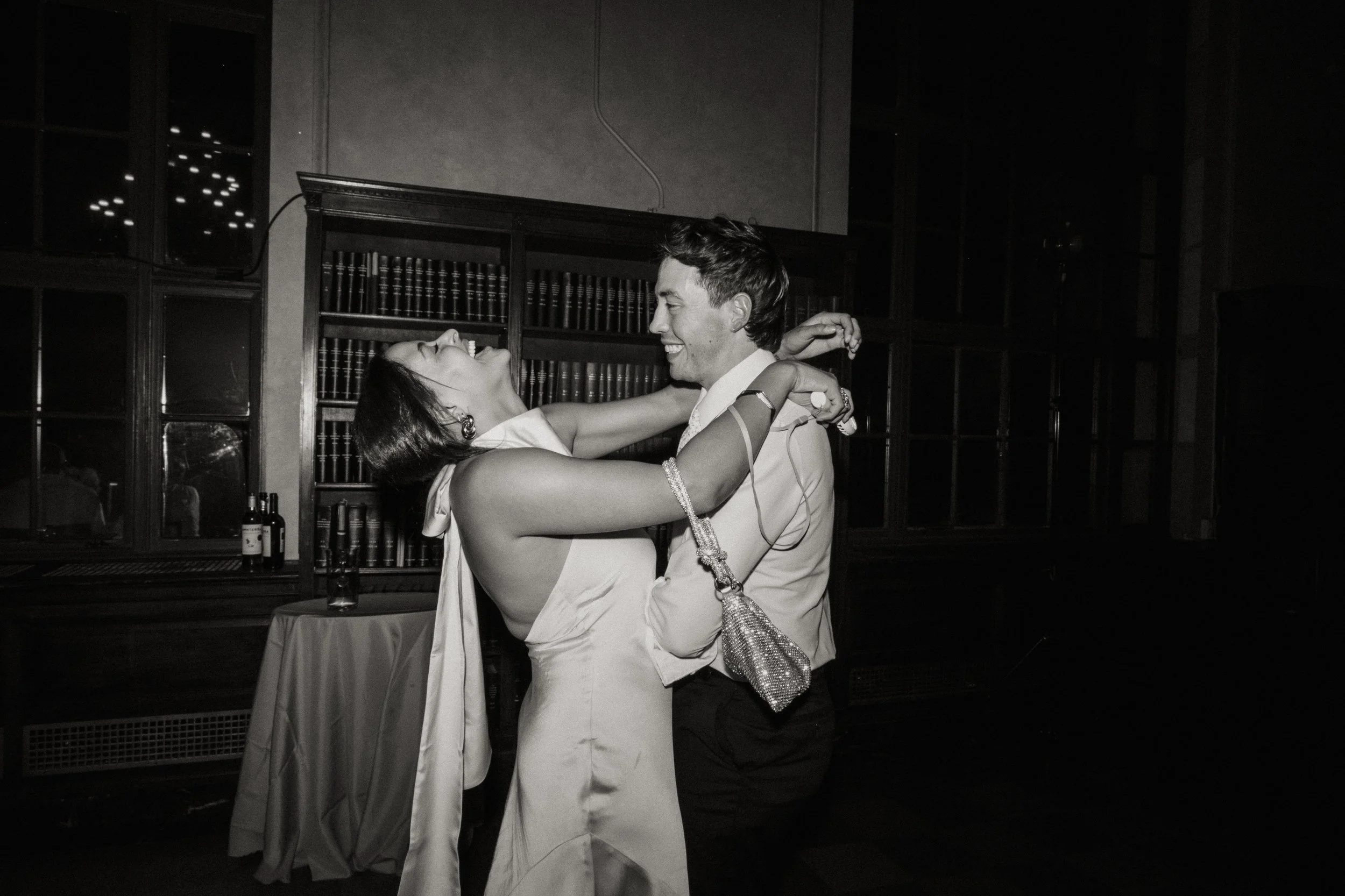 Black and white image of a man and woman dancing closely, smiling and main audience visible. The woman wears a sleeveless dress and holding a small purse, while the man wears a button-up shirt. They are in a room with dark wooden shelves, glass bottles, and a table.
