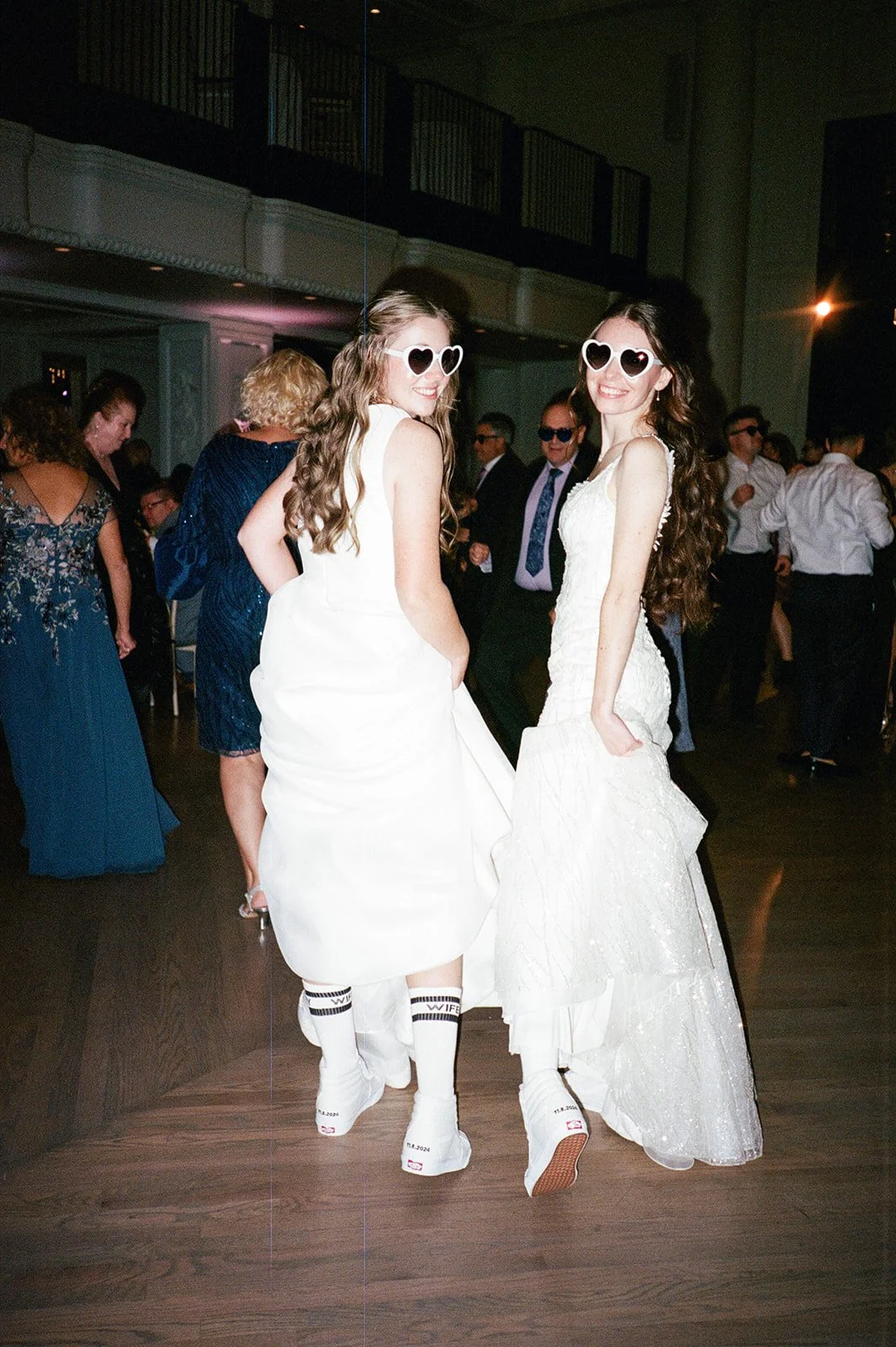 Two women in white dresses and heart-shaped sunglasses dancing at a wedding reception with guests in formal attire in the background.